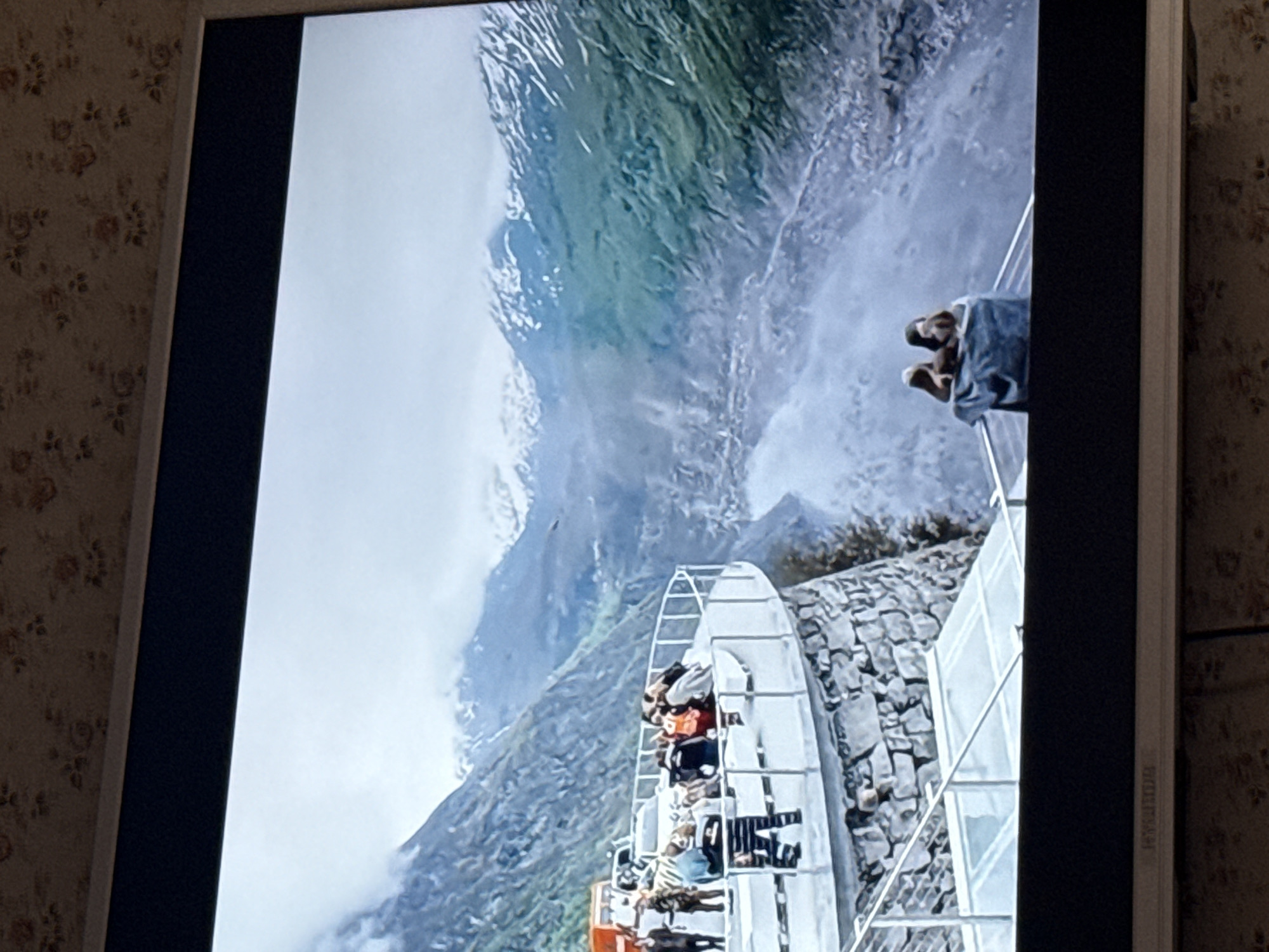 The image displays a television screen showing a mountainous landscape with a tourist attraction. The attraction appears to be a metal structure extending from the cliffside, containing multiple levels occupied by people. A waterfall is visible near the structure, descending from the mountain. Snow-capped peaks are present in the background, partially obscured by clouds. The surrounding environment includes stone walls and railings as part of the attraction. The TV screen is framed by a dark bezel and sits against a patterned wallpaper. There is no indication of the location being in Mytishchi, Russia.