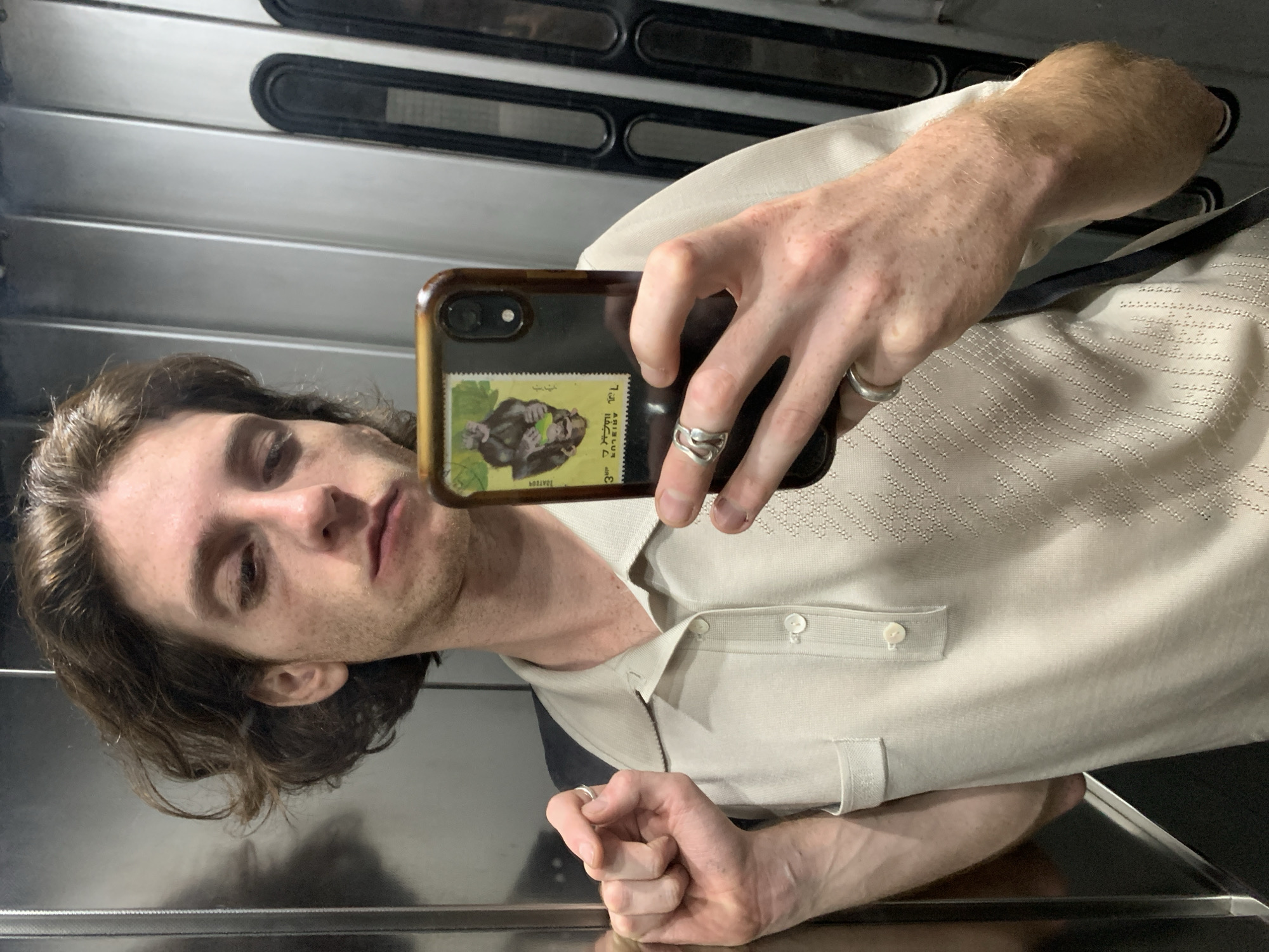 A young man with dark, wavy hair and fair skin is captured in a mirror selfie, likely in an elevator or a room with highly reflective, metallic wall panels. He gazes directly towards the smartphone held in his right hand, which has a dark-colored protective case. On the back of the phone case, a postage stamp-like sticker is visible, depicting a gorilla on a green and yellow background. The sticker features Arabic script, including the word "أبيجان" (Abidjan), and the numbers "٧" (7) and "3".
The man is wearing a light-beige or off-white polo shirt with a textured, patterned weave and a three-button placket. He has visible freckles on his skin and hands. On his left hand, he wears a simple silver band on his ring finger and a more ornate, wavy silver ring on his middle finger. A simple silver band is also on the ring finger of his right hand. The background consists of sleek, metallic horizontal panels with darker lines or vents at the top. His expression is neutral. The setting is indoors, with artificial lighting, consistent with a modern building in a city like Buenos Aires, Argentina, though no specific visual cues for the city are present.