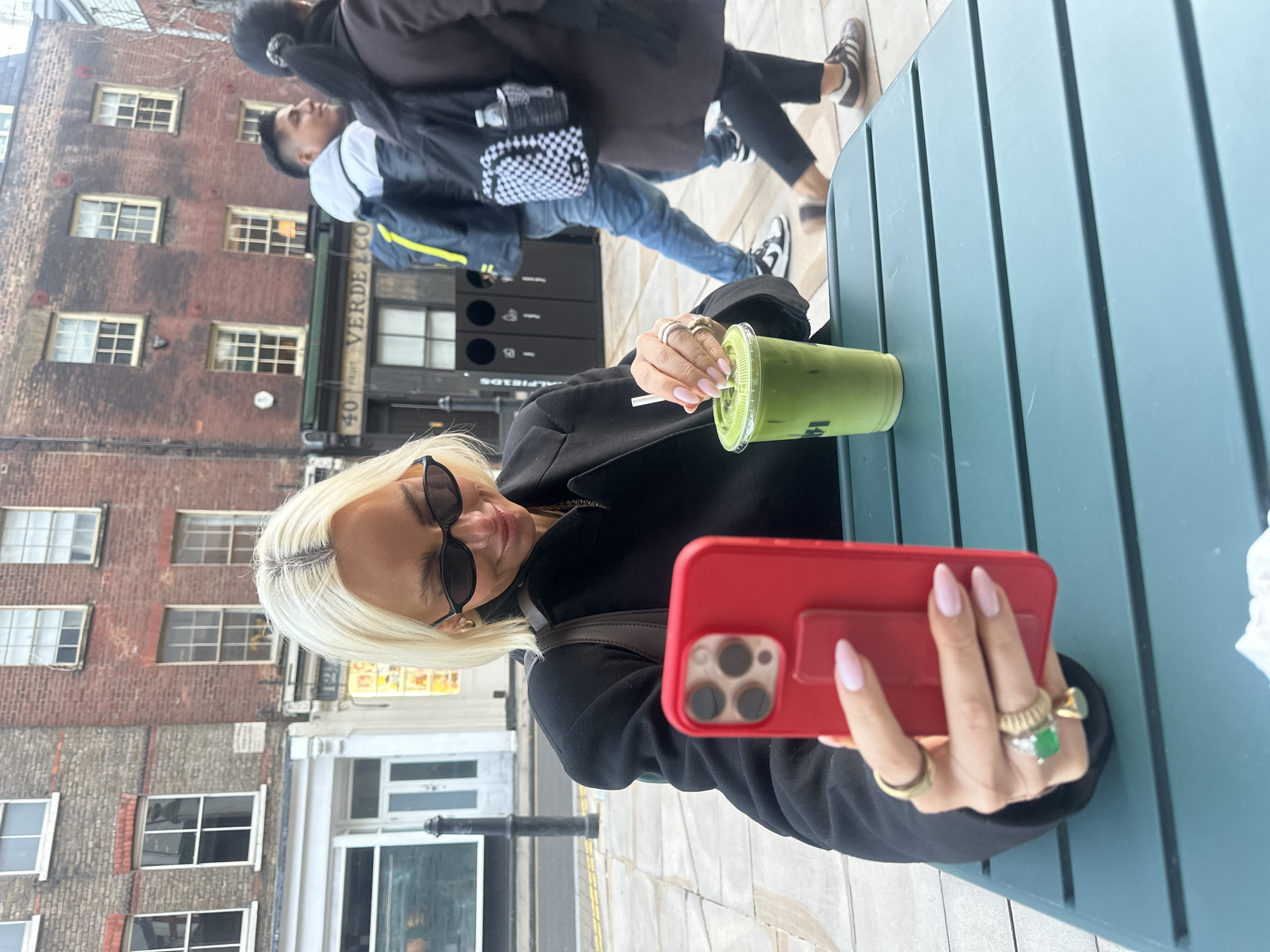 The scene depicts an outdoor cafe area on Brushfield Street, Spitalfields, London, United Kingdom, specifically in front of "Verde & Co" at address number 40. A fair-skinned woman with short blonde hair and black oval sunglasses is seated at a dark green slatted metal outdoor table. She wears a dark-colored blazer or coat and gold earrings. Her right hand, adorned with multiple gold rings including one featuring a large green stone, holds a red iPhone (likely a 15 Pro Max) with a red case and a matching pop socket-style grip, pointing it towards herself. Her fingernails are painted a light pink. A faint smile is visible on her face as she looks towards the phone screen. On the table to her left, a clear plastic cup contains a green iced beverage, likely a matcha latte, with a white straw. In the background, two pedestrians walk on a paved sidewalk from right to left. The foreground pedestrian is a male wearing a white and grey hooded top, blue jeans, and black and white sneakers, carrying a backpack with yellow accents. Behind him, a female in a dark coat and dark trousers wears Adidas-style sneakers and carries a black and white checkered backpack with a water bottle attached to the side. The street is lined with multi-story brick buildings featuring numerous white-framed sash windows. A black bollard is visible to the left, and three black recycling bins are positioned further back on the sidewalk, indicating public waste collection points. The overall environment suggests an urban setting on an overcast day.