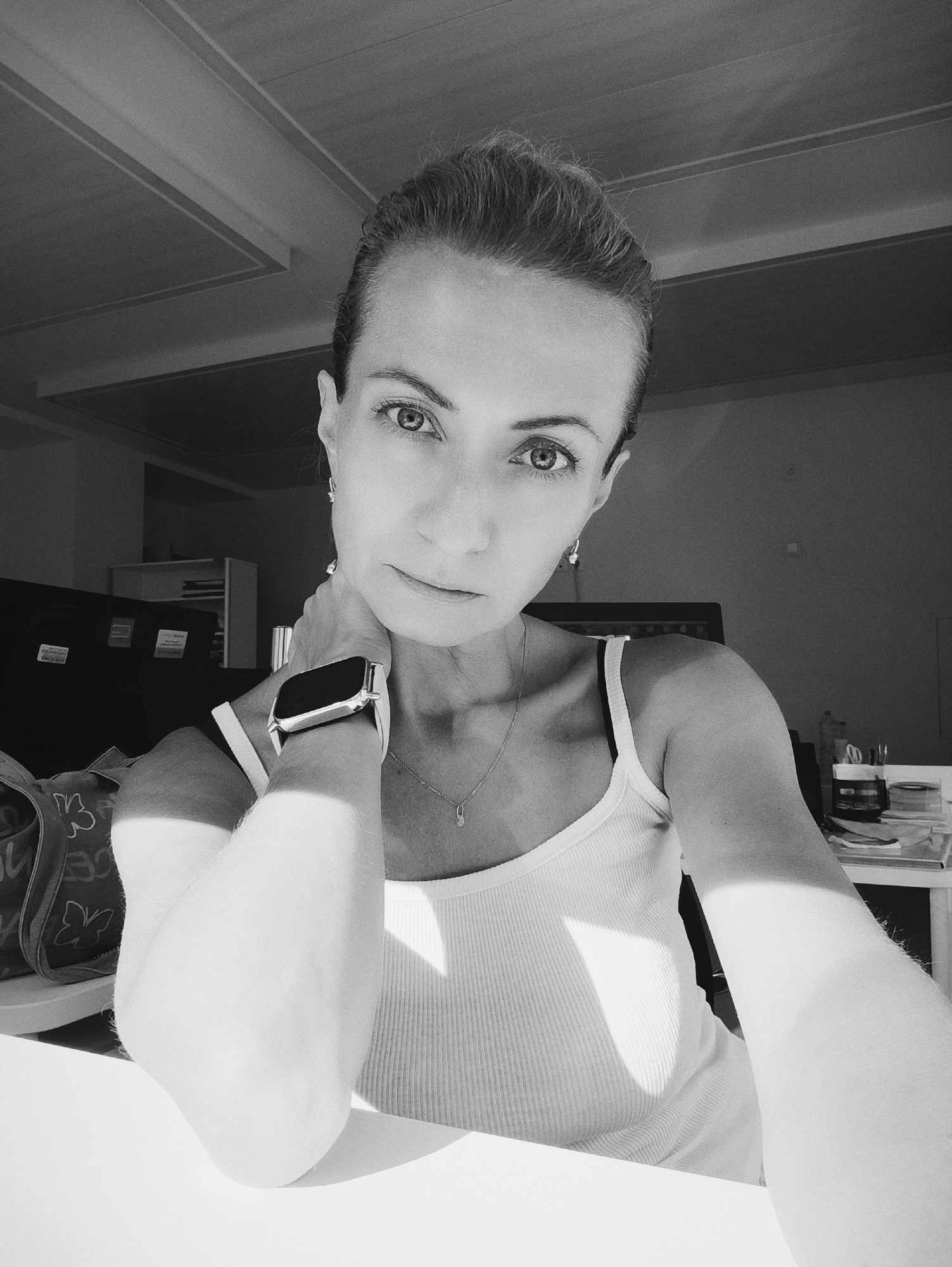 The image is a black and white, medium-close-up selfie of a woman taken indoors. She is looking directly at the camera with a neutral expression. She has her left arm bent, with her hand resting on the back of her neck. She is wearing a white, ribbed tank top, a necklace with a small pendant, and earrings. On her left wrist, she is wearing a smartwatch with a black face. Her hair is pulled back. Sunlight is casting shadows across her face and body. In the background, out of focus, are office-like objects including a desk, shelves, and what appears to be a computer monitor. There is no visible indication that this image was taken in Moscow, Russia.