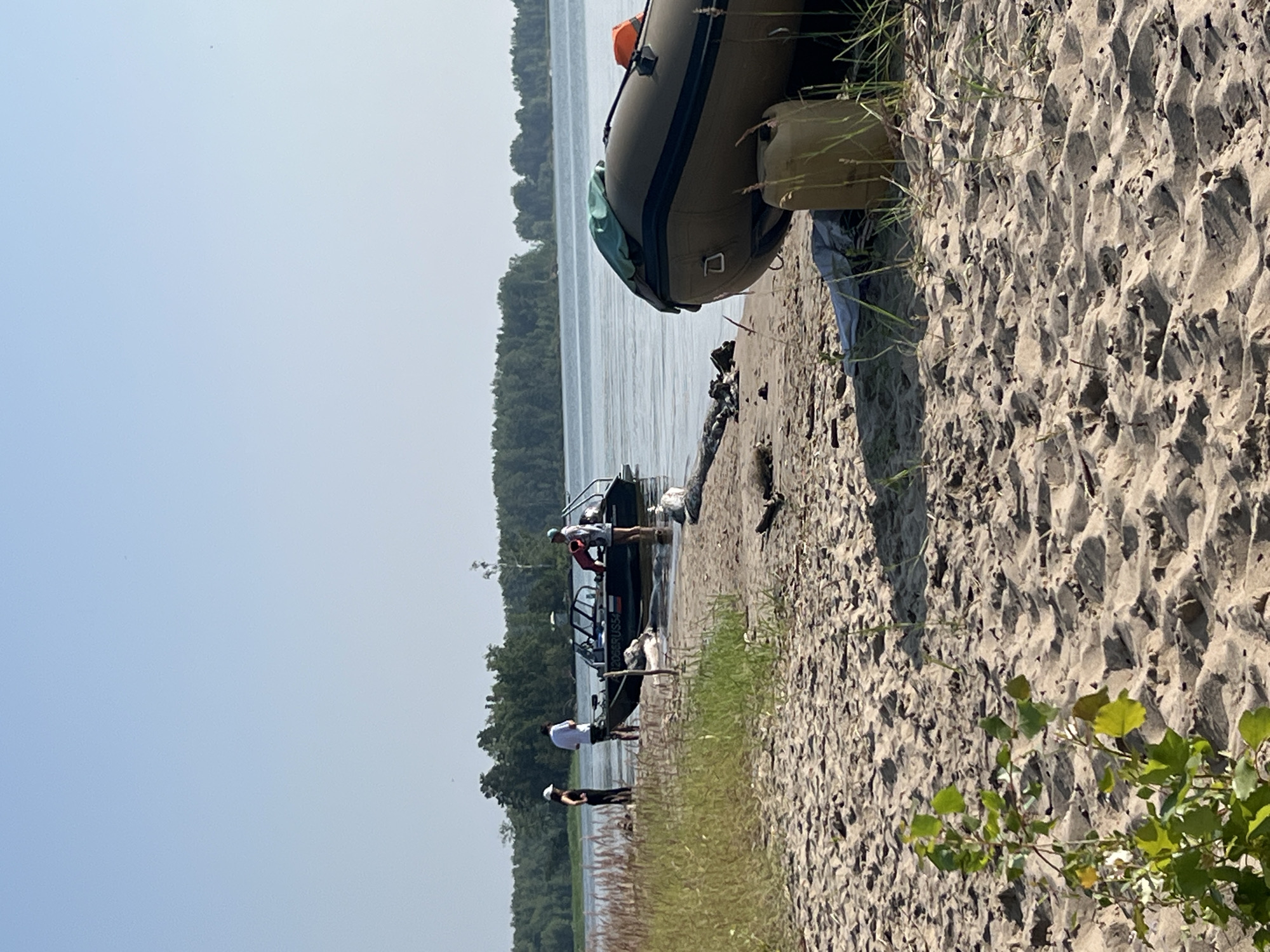 A daytime, eye-level, medium shot shows a sandy beach on a clear day, with a body of calm water stretching to a tree line in the distance. In the foreground, a large, olive-green inflatable boat rests on the sand, partially covered with a green tarp, with an orange life vest visible on its rear. Next to the boat, a tan plastic container is placed on the sand. A smaller, dark-colored motorboat is pulled partially onto the shore in the mid-ground. Three individuals are visible near the motorboat; two are standing on the sand, and one is partially submerged in the water. The person on the left is a woman wearing a white hat and a dark top and pants. The person in the middle, also standing on the sand, is a man in a white t-shirt and dark shorts. The third person, near the motorboat, appears to be a man in a light-colored shirt and shorts, standing in the water. The sand is textured with footprints and scattered driftwood. The background features a dense forest bordering the water, under a clear, pale blue sky.
