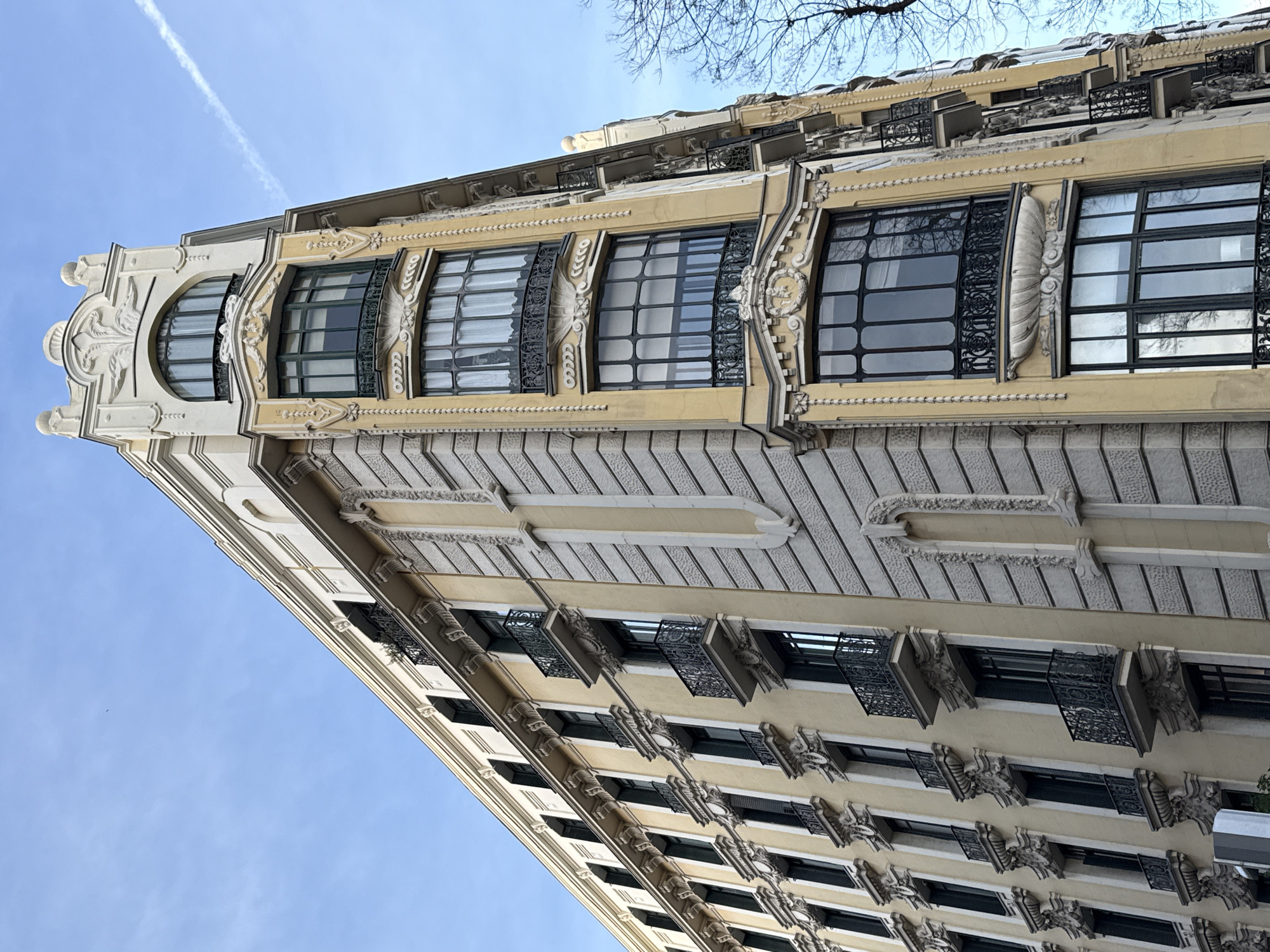 The image shows a low-angle view of a building in Madrid, Spain. The building has a distinctive triangular shape and is decorated in an ornate, classical style. The building features a facade with a mix of stone and plaster, with multiple floors of windows framed by decorative ironwork and carved stone elements. There is a blue sky with a faint contrail is in the background.