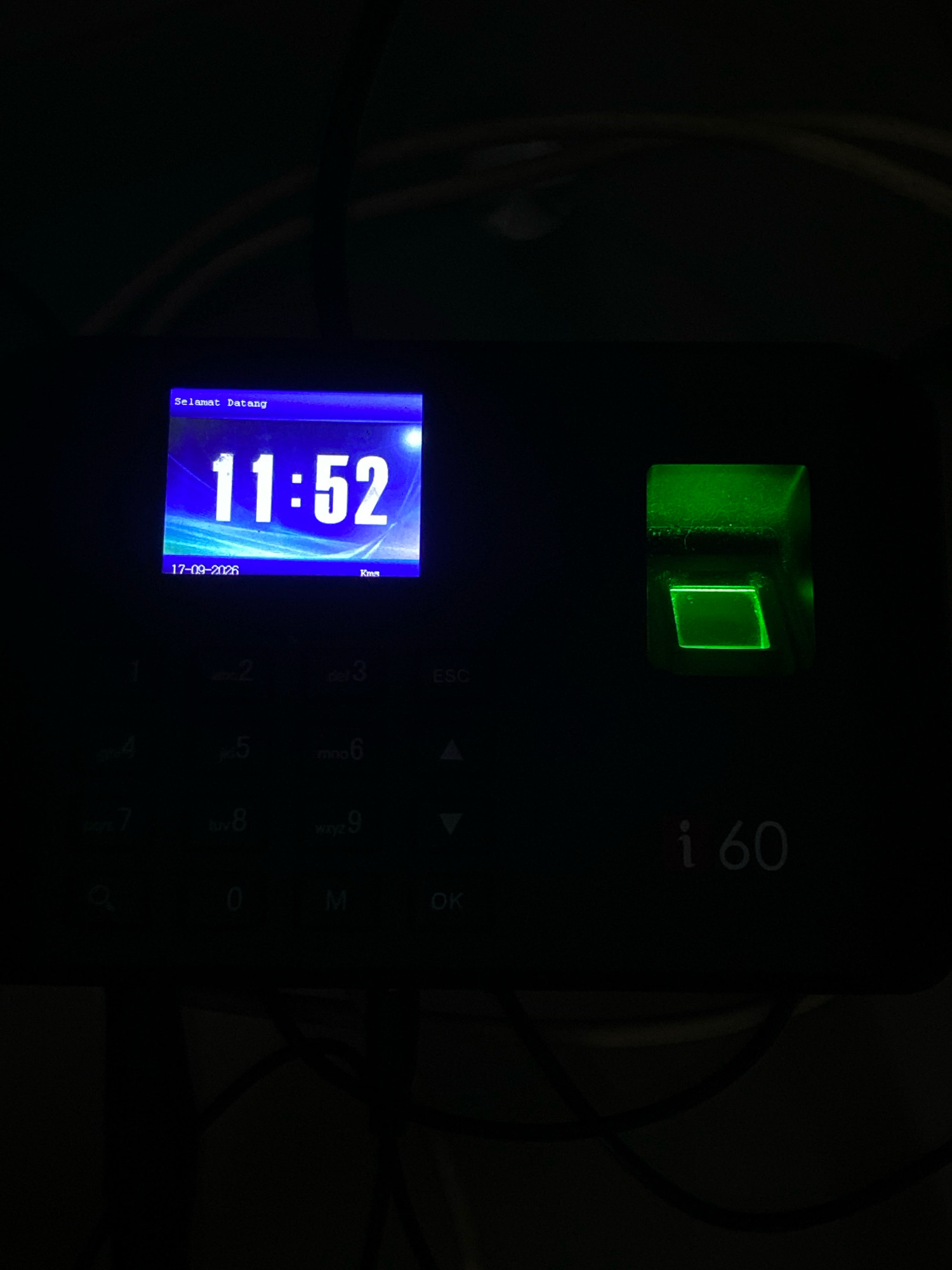 The image shows a close-up of a biometric time clock device. The device has a digital display showing the time as "11:52" and the date as "17-09-2026". The screen also displays the text "Selamat Datang" in a blue hue. To the right of the screen, there is a green illuminated fingerprint scanner. Below the screen is a keypad with numbers and letters, as well as buttons labeled "ESC", "M", and "OK". The text "i60" is visible in the lower right corner of the device. The device is black and appears to be situated in a dimly lit environment, possibly indoors. The context of "Klurak, Indonesia" is not visually represented within the image itself.