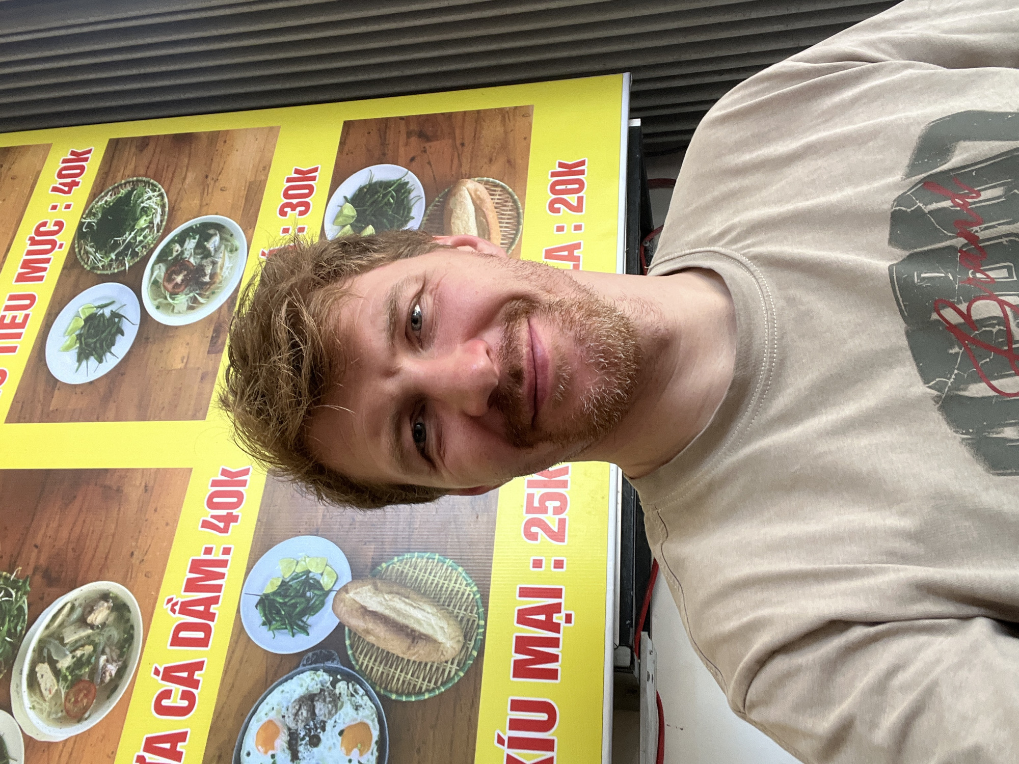 An indoor head-and-shoulders portrait captures an adult male, approximately 25-35 years old, with fair skin, short reddish-brown hair, and a neatly trimmed beard. He exhibits a slight smile, gazing directly forward. He is dressed in a light beige crew-neck t-shirt, featuring a partially visible dark graphic design on the chest.
The background consists primarily of a bright yellow commercial banner, likely a menu, adorned with red Vietnamese text and images of various food items. Visible text includes phrases like "CÁ DẦM: 40k", "TIÊU MỰC : 40k", "MẠI : 25k", and "A : 20k," with 'k' representing thousands of currency units. Depicted dishes include bowls of soup with vegetables and sliced tomatoes, fried eggs with a base, baguettes in woven baskets, and plates of green vegetables with lime. Beneath the banner, red electrical wiring is visible. A dark, vertically ribbed wall section occupies the right side of the frame. The scene is well-lit. The location is a food establishment in Unknown, Unknown.