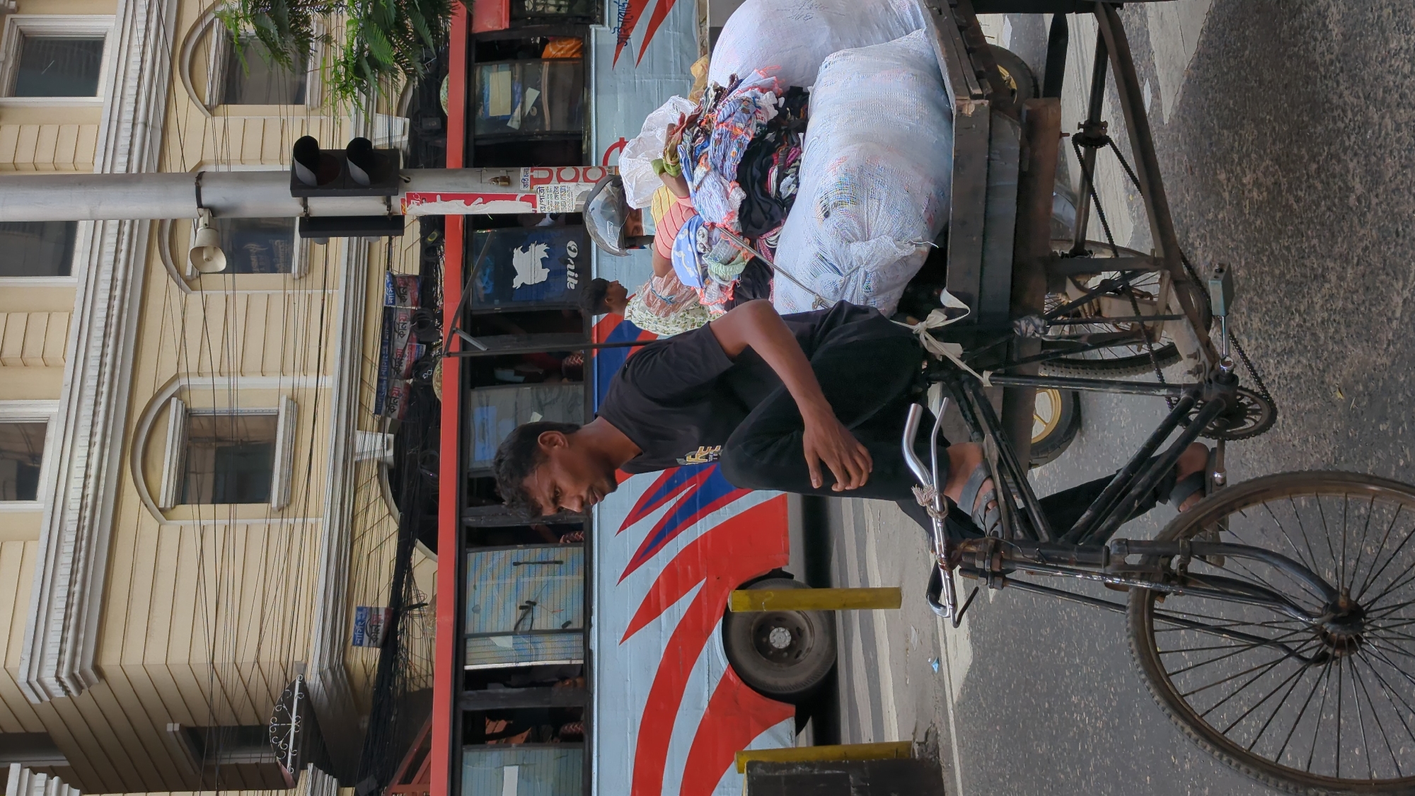 A man is riding a bicycle rickshaw with a large load of sacks filled with clothing. The bicycle rickshaw is facing the right of the frame. The man is dark-skinned, wearing a dark shirt and pants. A bus with a red and blue stripe pattern is visible behind him. A light-colored building with multiple windows stands on the left side of the frame. The presence of overhead electrical wires is evident. This is likely a street scene in Dhaka, Bangladesh.