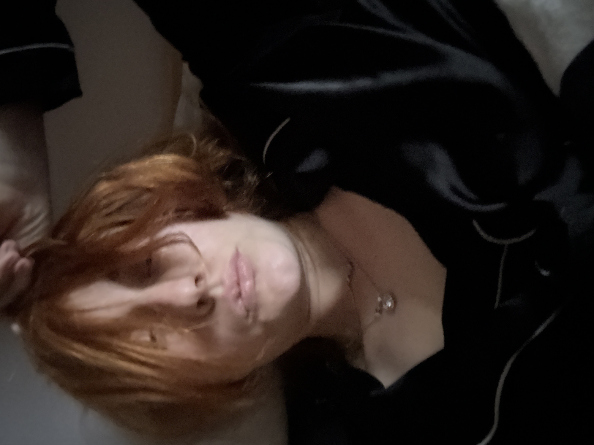 A woman with red hair is lying down on a white surface, possibly a bed. She is wearing a black silky pajama set with white piping. A silver necklace with a circular pendant is visible on her neck. Her lips are slightly puckered. The scene's location is not discernible from the image.