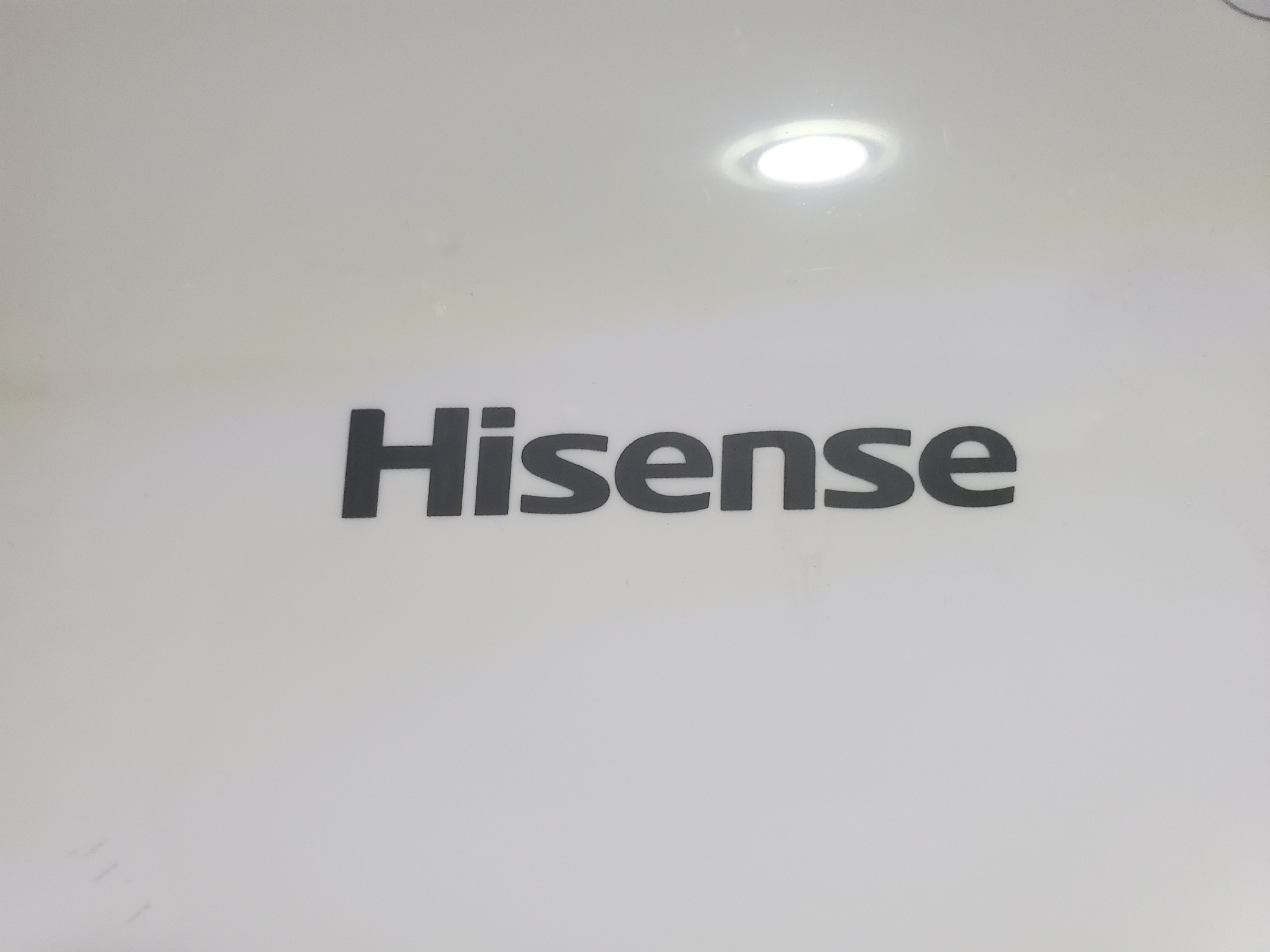 A Hisense logo is displayed in black on a white background. The logo is centrally located on the surface, which appears to be a wall or appliance. Above the logo, a circular light fixture is visible, casting a soft glow. The overall lighting is bright and even, with no significant shadows. The image is clear and sharp, with the text easily readable.