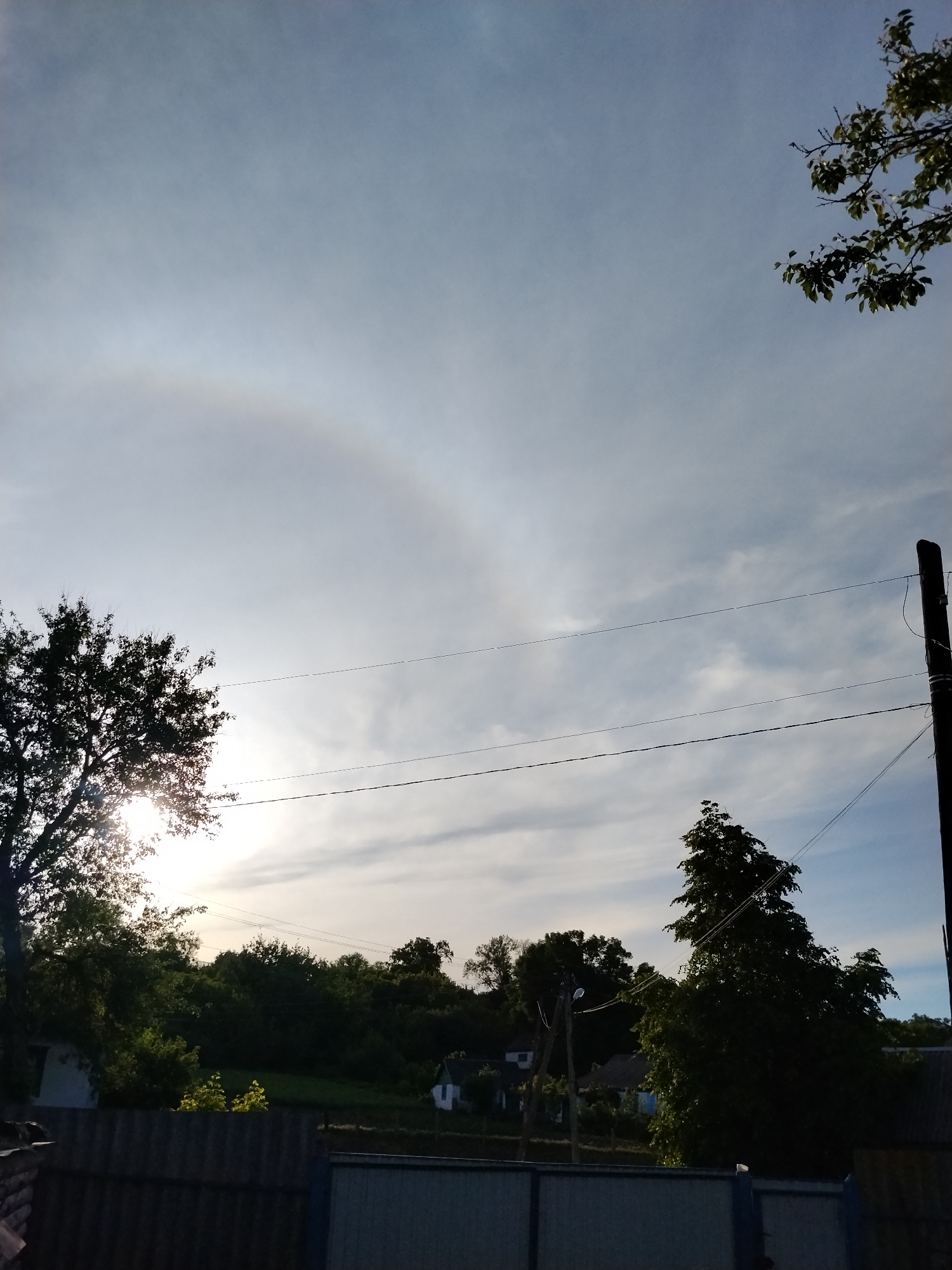 The image captures a rural or semi-rural landscape under a bright, partly cloudy sky, with a visible solar halo. The sun, partially obscured by the foliage of a large tree on the left, casts a strong flare into the camera lens. Above the sun, a distinct, faint, circular rainbow-like halo is visible in the clouds, arcing across the upper left sky.
In the foreground, the top of a dark, corrugated fence stretches across the bottom left, meeting a lighter, slatted fence that extends across the bottom right. Lush green trees and foliage dominate the midground, forming a dense line along the horizon. Among these trees, small, light-colored houses or buildings are discernible, suggesting a settlement beyond the fences.
On the far right, a dark utility pole stands tall, with two thin power lines extending horizontally across the mid-sky towards the left, disappearing behind the tree that partially blocks the sun. Another branched tree is visible in the upper right corner, with its leaves silhouetted against the sky.
The time of day appears to be late afternoon or early morning due to the low angle of the sun and the soft, diffused light, which creates some silhouetting of foreground elements. The weather is bright with scattered clouds, conducive to the formation of the observed sun halo. The scene evokes a peaceful, natural setting, likely in the Ukrainian countryside. There is no human activity or visible text in the image.