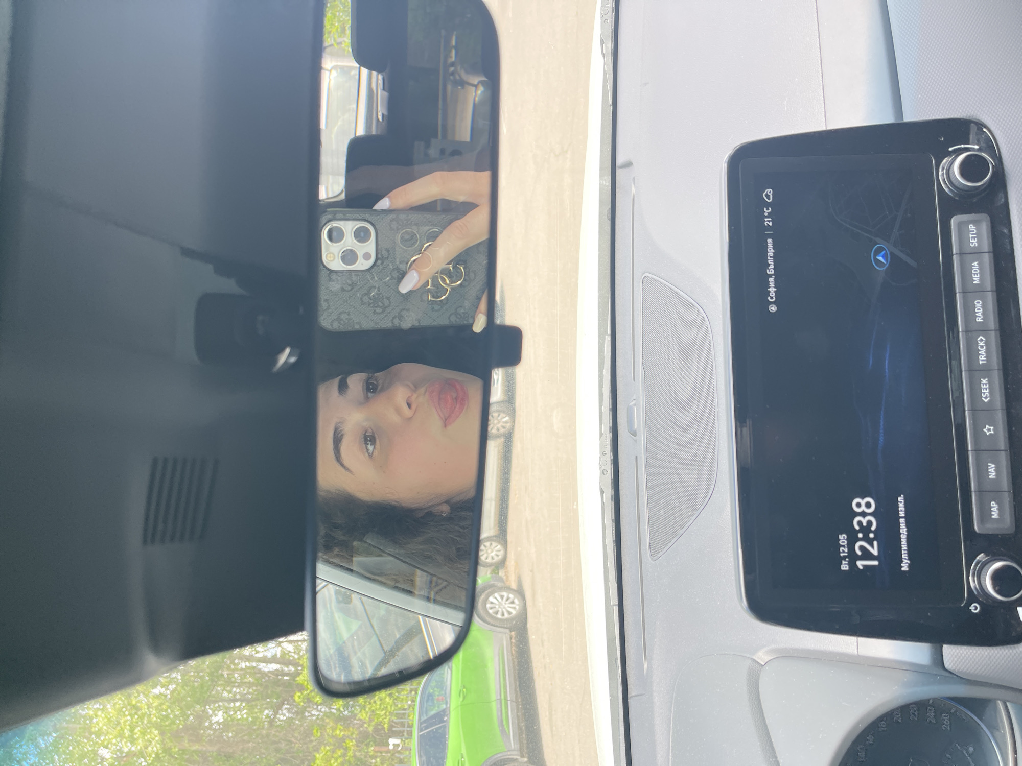 The image shows a selfie taken in the rearview mirror of a car. A woman is visible in the mirror, holding up a phone to take the picture. The car's dashboard is visible to the right of the mirror, displaying "София, България | 21°С" indicating the location is Sofia, Bulgaria and the temperature is 21°C. The time displayed on the dashboard is 12:38 on Tuesday, May 12th. A green vehicle is partially visible in the mirror's reflection. The image suggests the person is either in or near a vehicle in Sofia, Bulgaria.
