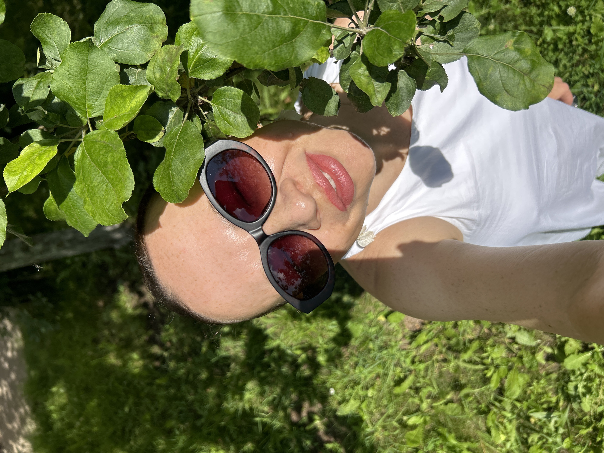 A woman with fair skin and dark hair, wearing black sunglasses and a white dress, is outdoors in a grassy area with sunlight filtering through green leaves. She is looking up and smiling.