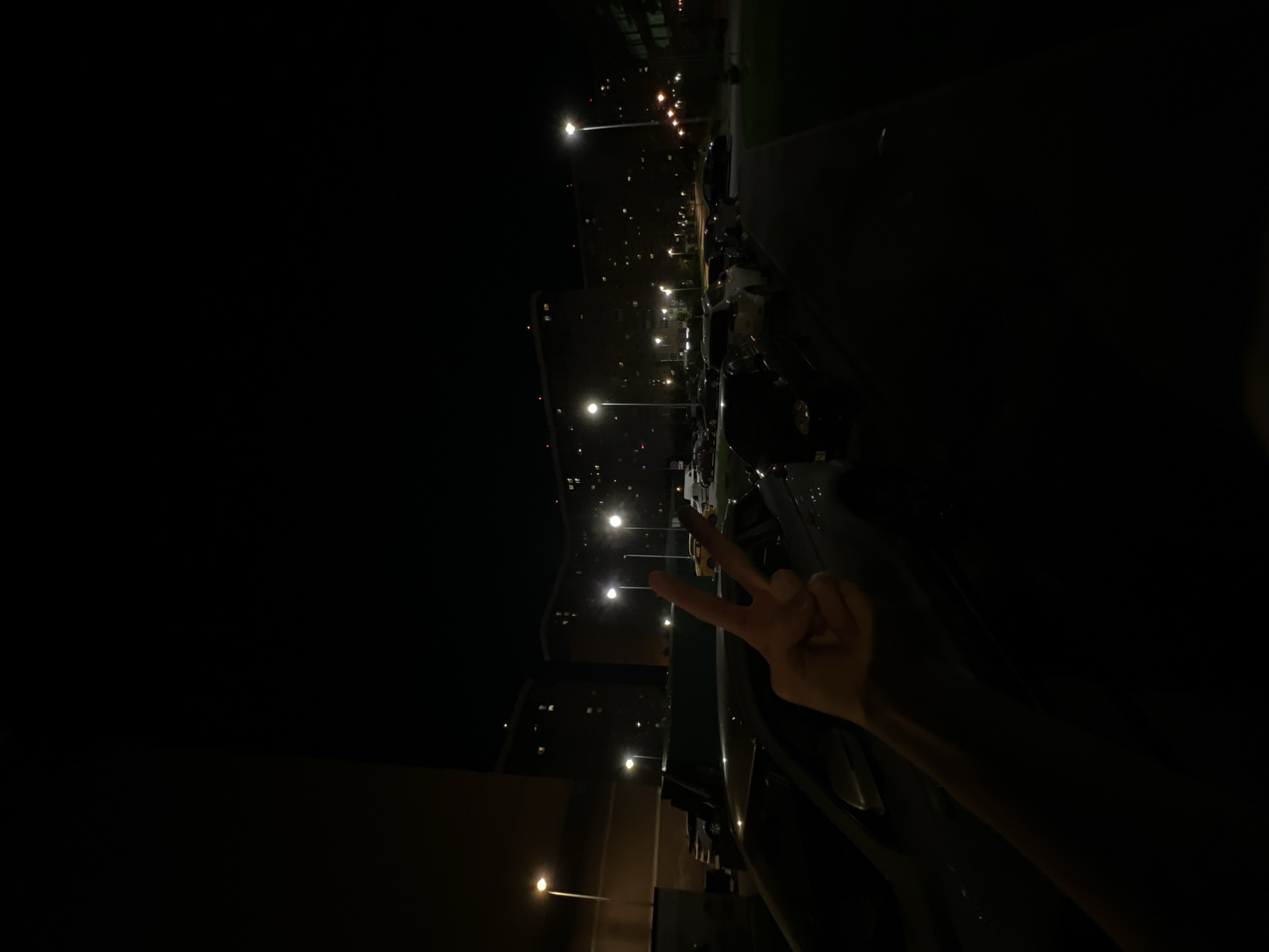 A dark, rotated night shot shows an arm and hand making a peace sign near a car, with apartment buildings, streetlights, and parked vehicles in the background. The scene appears to be located in Lyubertsy, Russia.