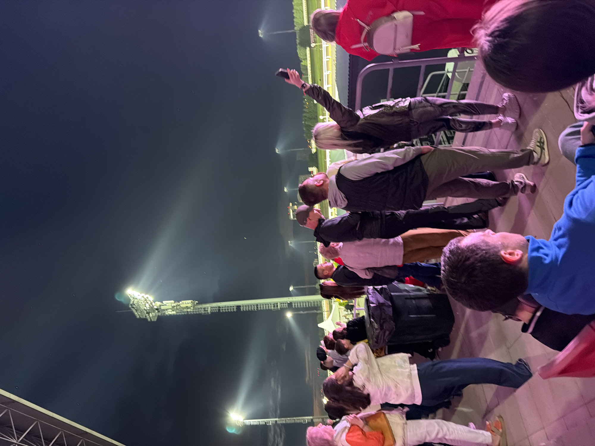 A group of people stand on a walkway at night, illuminated by stadium lights. The foreground shows a man in a blue jacket, his back to the camera. To his left is a red seat. Behind him, a crowd of people are standing and looking towards a bright light tower in the distance. Some are holding phones up, possibly taking pictures or videos. The ground is paved with pinkish lighting. In the background, a race track or similar venue is visible, bordered by lights and trees. A large black trash bin is positioned in the middle ground.
