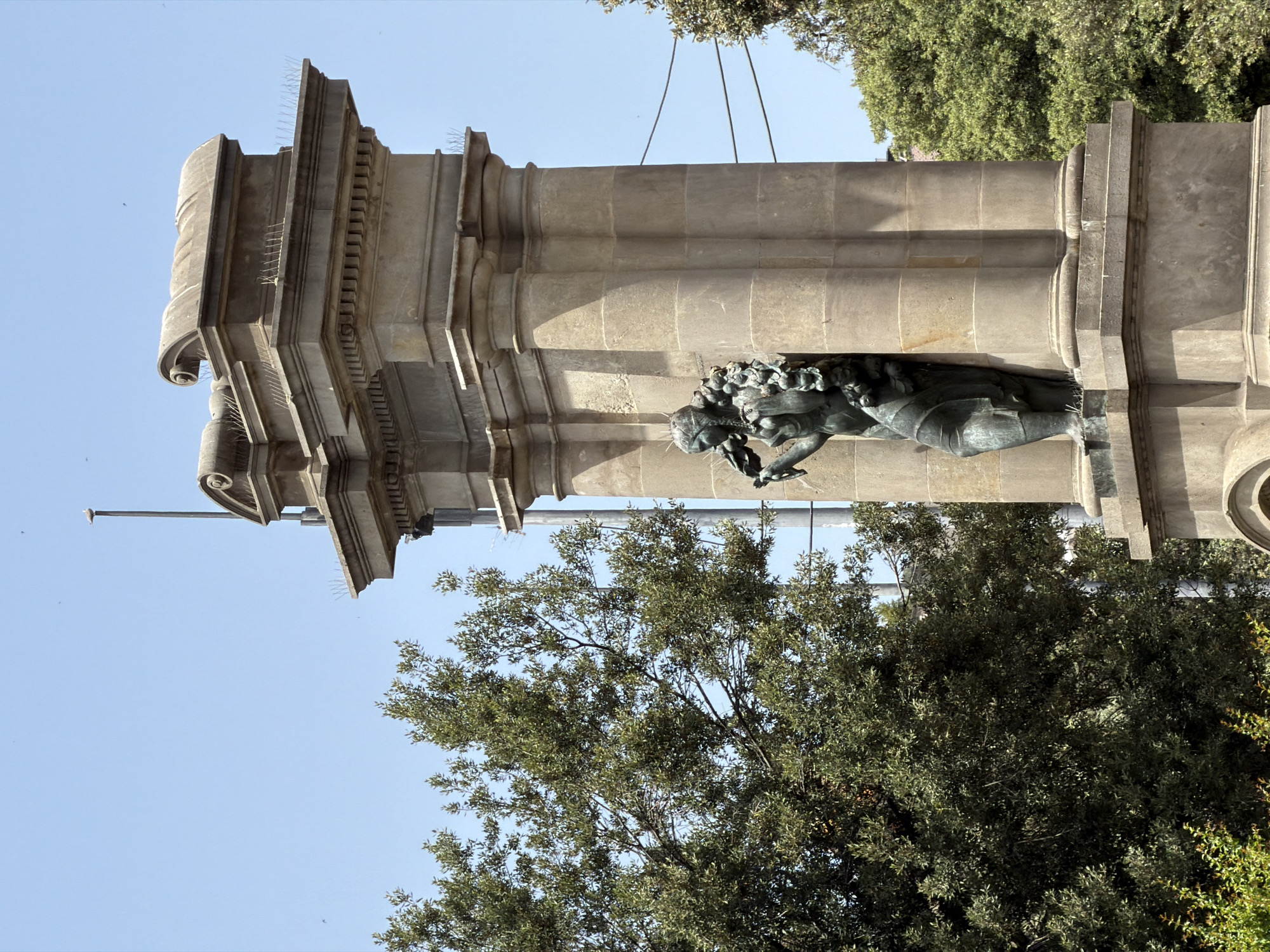 This is a low-angle, vertical photograph of a stone monument with a bronze sculpture embedded in its side. The monument is tall and made of large, beige stone blocks with ornate carvings and decorative elements, including what appear to be stylized columns. A bronze sculpture of a winged figure, possibly an angel or a mythical creature, is visible lying down, holding a wreath or floral arrangement. The sculpture has a weathered, green patina.
The monument is situated outdoors, with a clear blue sky in the background. Lush green trees with dense foliage fill the lower part of the frame and frame the right side of the monument. The sun appears to be shining, casting shadows and highlighting the textures of the stone and sculpture, suggesting it is daytime. Several small, dark specks that might be birds or insects are scattered across the sky.
The overall scene evokes a sense of history and artistry, likely in a public space such as a park or plaza. The presence of the trees and the clear sky suggest pleasant weather. No text is visible in the image. The location is identified as Barcelona, Spain.