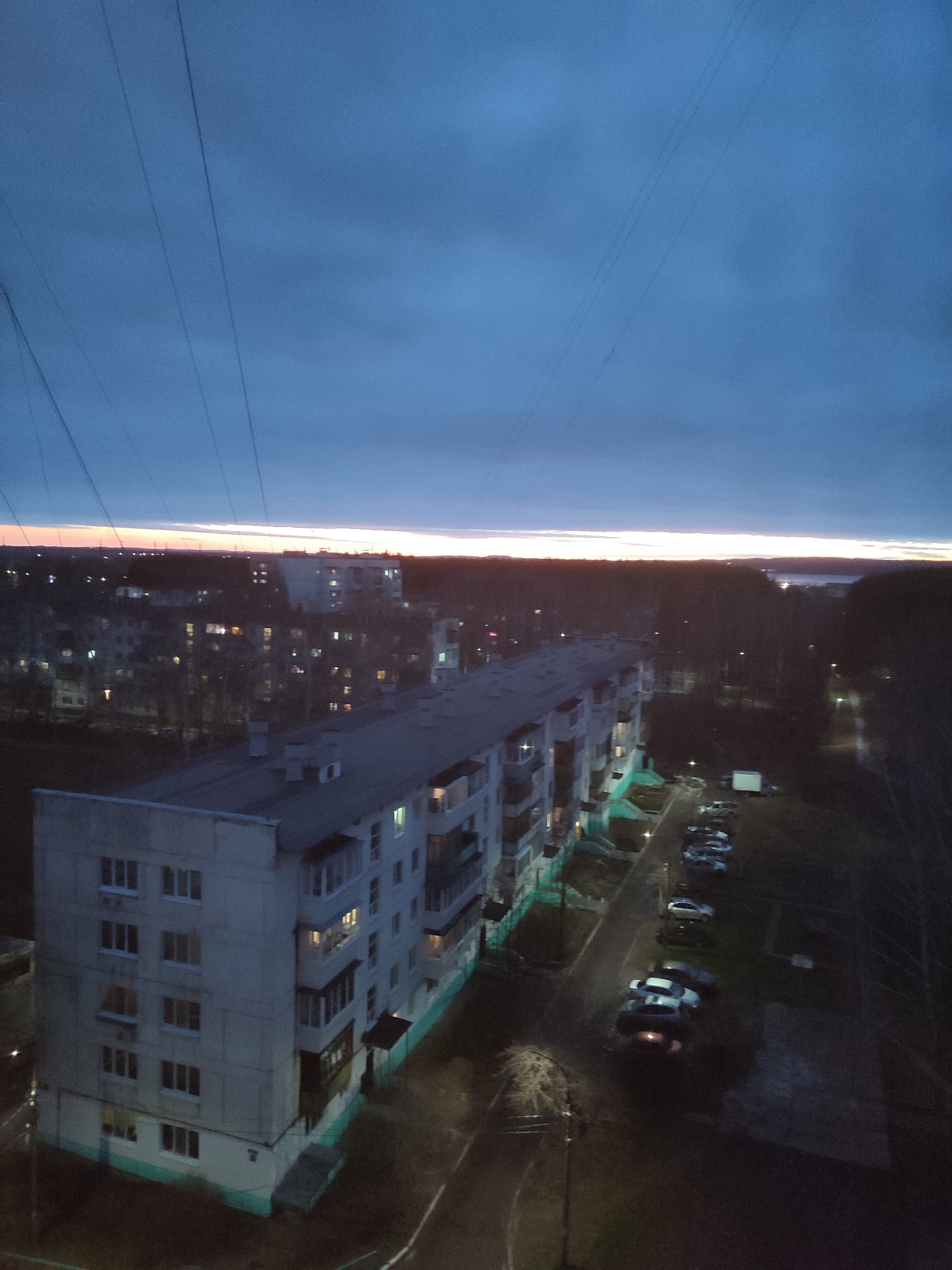 The image presents a high-angle view of an urban or suburban landscape during twilight, likely dusk, characterized by a deep blue sky with a vibrant orange and yellow glow along the distant horizon. Overhead, several thin, dark wires or power lines traverse the blue sky.
The primary subject is a long, multi-story residential building, possibly an apartment complex, occupying the mid-ground and lower left of the frame. This building has a grey facade, numerous windows (many illuminated), and several balconies. A strip of teal or light green paint runs along its base. Adjacent to the building, a paved area serves as a parking lot, accommodating several parked cars. Visible streetlights emit a soft glow onto the ground.
Beyond this main structure, the background reveals a sprawling expanse of other buildings with lit windows, interspersed with dark, bare trees, suggesting a cooler season. In the far distance, a body of water may be discernible on the right side of the horizon, reflecting the faint light. The overall scene is devoid of visible people or direct activity, conveying a sense of quietude as artificial lights begin to dominate the receding daylight. No discernible text is visible in the image.