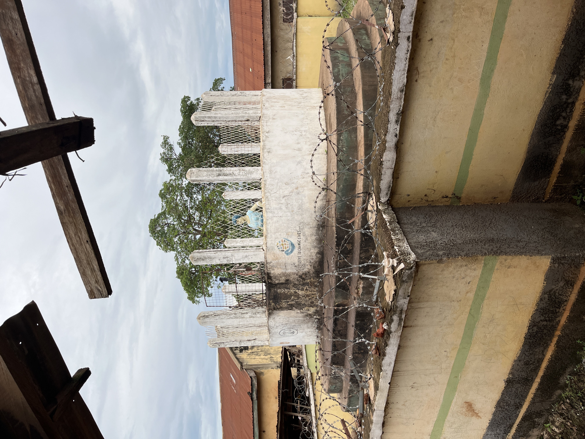 This image captures an outdoor scene in Idah, Nigeria, under an overcast sky. The central subject is a multi-tiered, circular concrete monument. The uppermost tier is a white cylindrical structure, approximately 1.5 meters in height, supported by five visible concrete pillars that enclose a metal mesh fence. On the front-facing surface of the white cylinder, a blue globe emblem depicting two hands, characteristic of Rotary International, is visible to the right. Below this emblem, partially obscured text reads, "SERVE TO CHANGE LIVES..." A light blue mannequin or statue is positioned behind the metal mesh on the right side of the cylinder.
The monument's lower tiers consist of dark grey concrete steps, each heavily adorned with coils of concertina wire. The entire concrete structure shows signs of weathering, dirt, and biological growth. In the immediate foreground, a concrete retaining wall, painted yellow with a distinct horizontal green stripe across its middle and a darker grey lower section, is also topped with concertina wire and scattered debris including broken red bricks and fabric scraps. The ground directly in front of this wall consists of sparse earth.
In the background, two single-story buildings are visible. The building on the left features a rusty, corrugated metal roof and a faded yellow wall, with a darker, open area beneath an extended overhang. The building on the right also has a rusty, corrugated metal roof and yellow walls. Dense green foliage from large trees is visible behind these structures and the monument.
Two rough-hewn wooden beams are prominent in the upper portion of the frame, suggesting the viewpoint is from within or beneath a structure with an open or damaged roof. No active human figures are depicted in the scene.