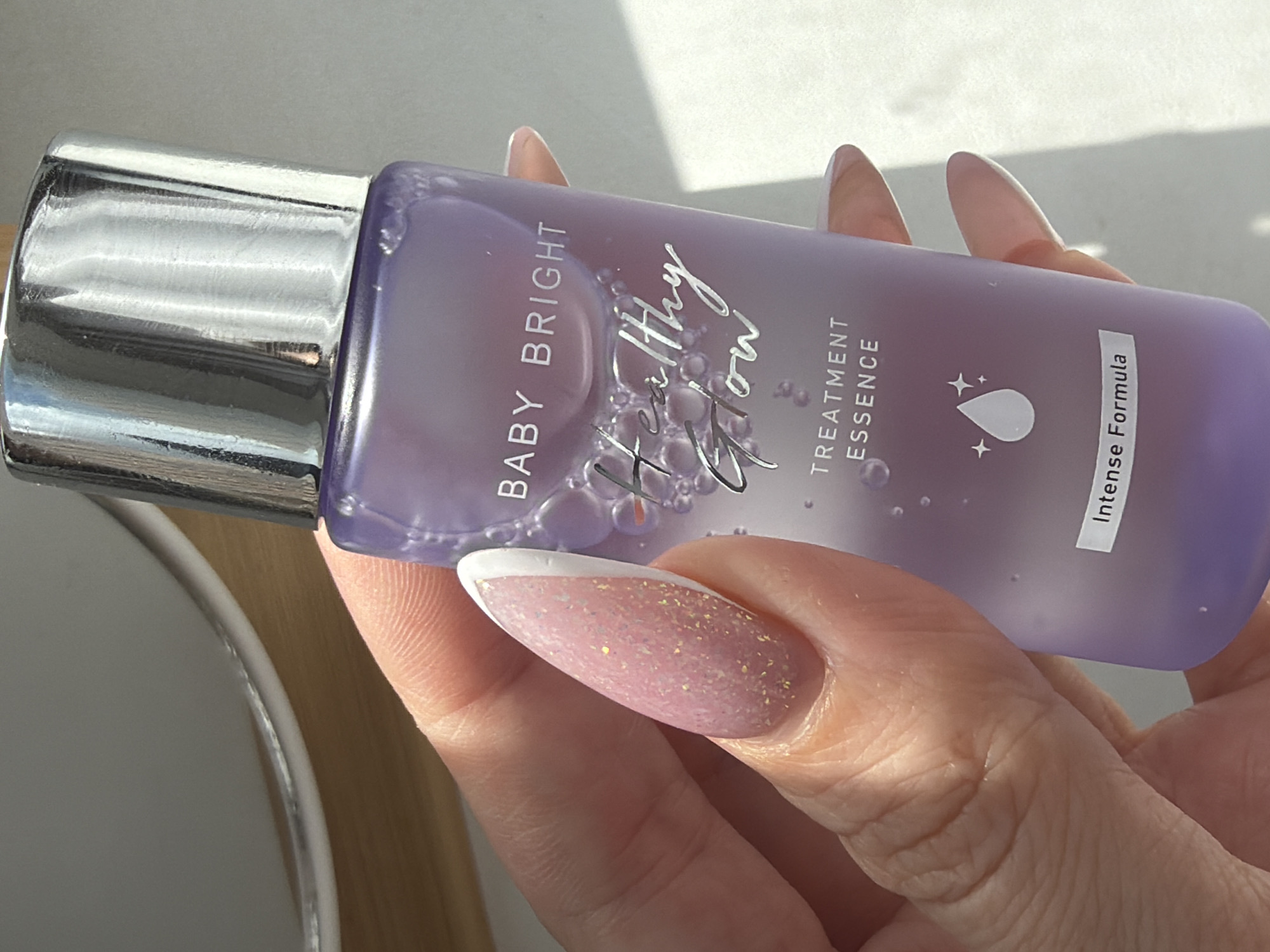 A light-skinned hand holds a bottle of "Baby Bright Healthy Glow Treatment Essence." The bottle is purple with a silver cap. The fingernails are long and oval, with a pink glitter base and a white French tip. The background is blurry and indistinct but appears to contain parts of a white sink and a wooden surface. The image does not provide any information about location in the city of Saint Petersburg, Russia.