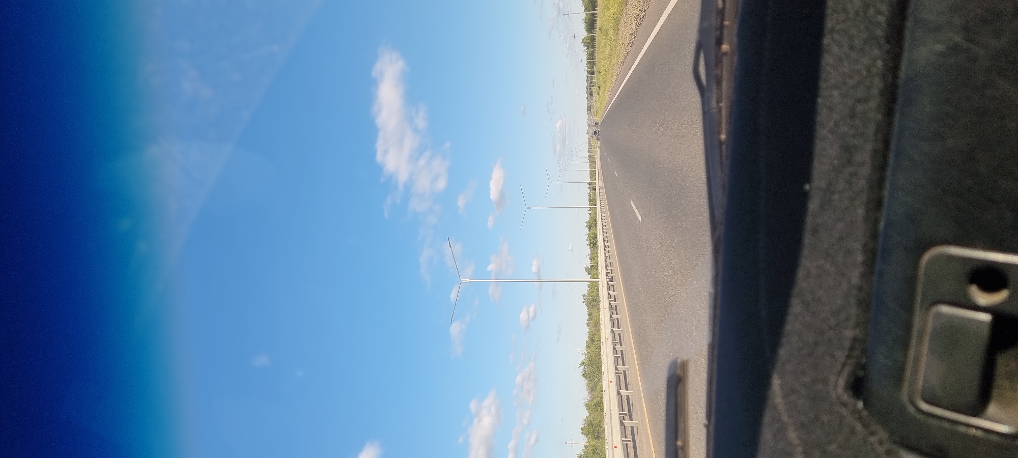 The image captures a view from inside a vehicle traveling on a highway. The road ahead is paved and has lane markings. On the left side of the road, there is a guardrail and a line of trees and vegetation. Several streetlights are visible along the roadside, spaced at regular intervals. The sky is blue with scattered white clouds. In the distance, a few other vehicles can be seen on the highway. The foreground shows the dashboard of the vehicle, including part of the steering wheel and windshield wipers. The location is identified as Bestamak, Kazakhstan. The scene depicts a typical highway environment on a clear day.