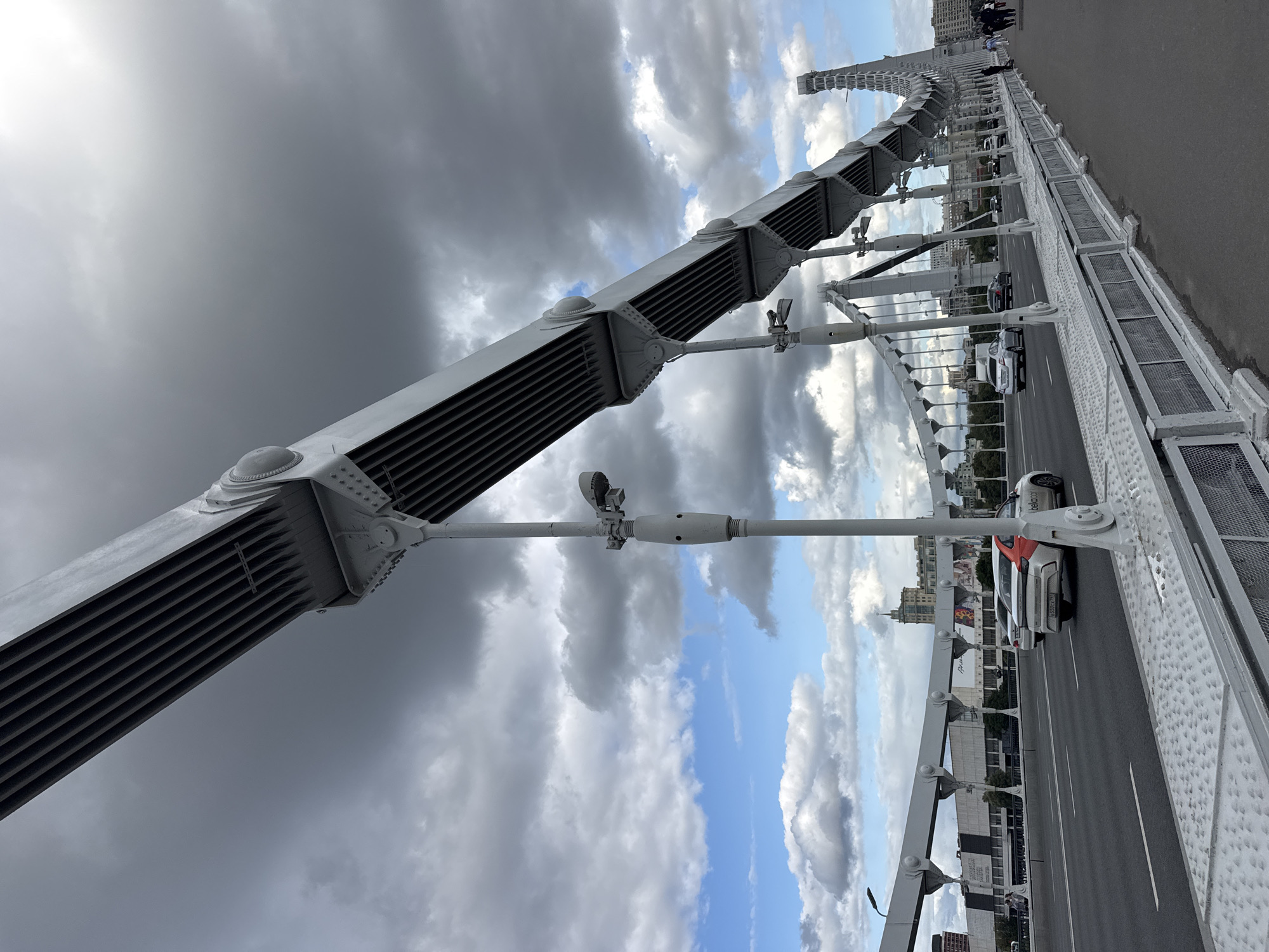 The image shows a view of the Krymsky Bridge in Moscow, Russia, on a cloudy day. The bridge is a suspension bridge with a distinctive white metal structure. The foreground is dominated by the bridge's infrastructure, including large metal beams, cables, and lampposts. Cars are seen driving on the roadway of the bridge, and pedestrians are walking on the sidewalk. In the background, there are buildings and a cityscape, typical of Moscow. The sky is filled with dramatic, dark clouds interspersed with patches of blue sky and sunlight.
