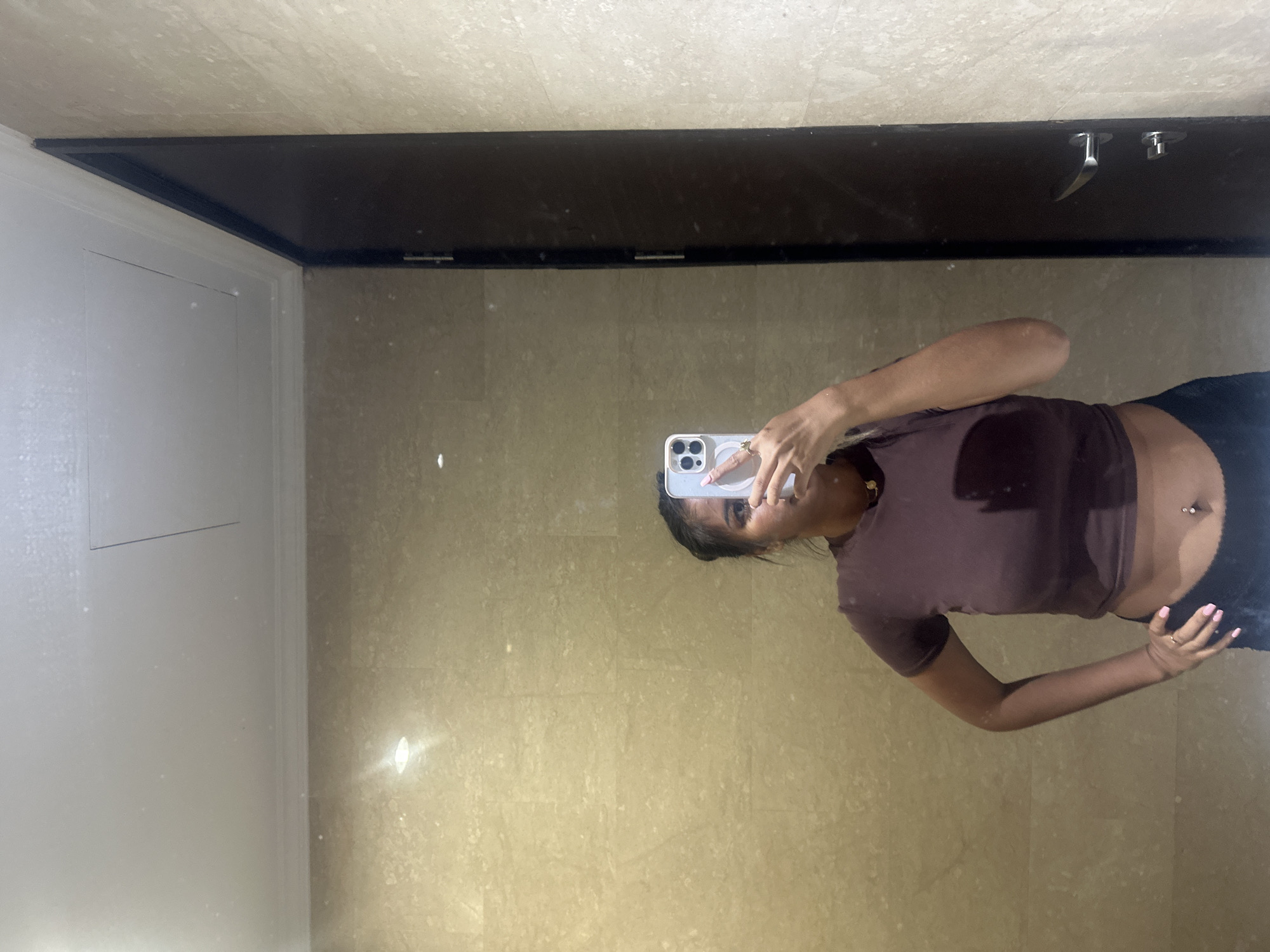 A person is taking a mirror selfie in what appears to be a bathroom. The person is wearing a brown crop top and dark jeans or pants. The bathroom has beige tile walls and a dark, rectangular mirror with chrome faucet fixtures. There is a white door or panel to the left of the mirror. The location is not discernible from the image, and cannot be specifically identified as Central Jakarta, Indonesia.