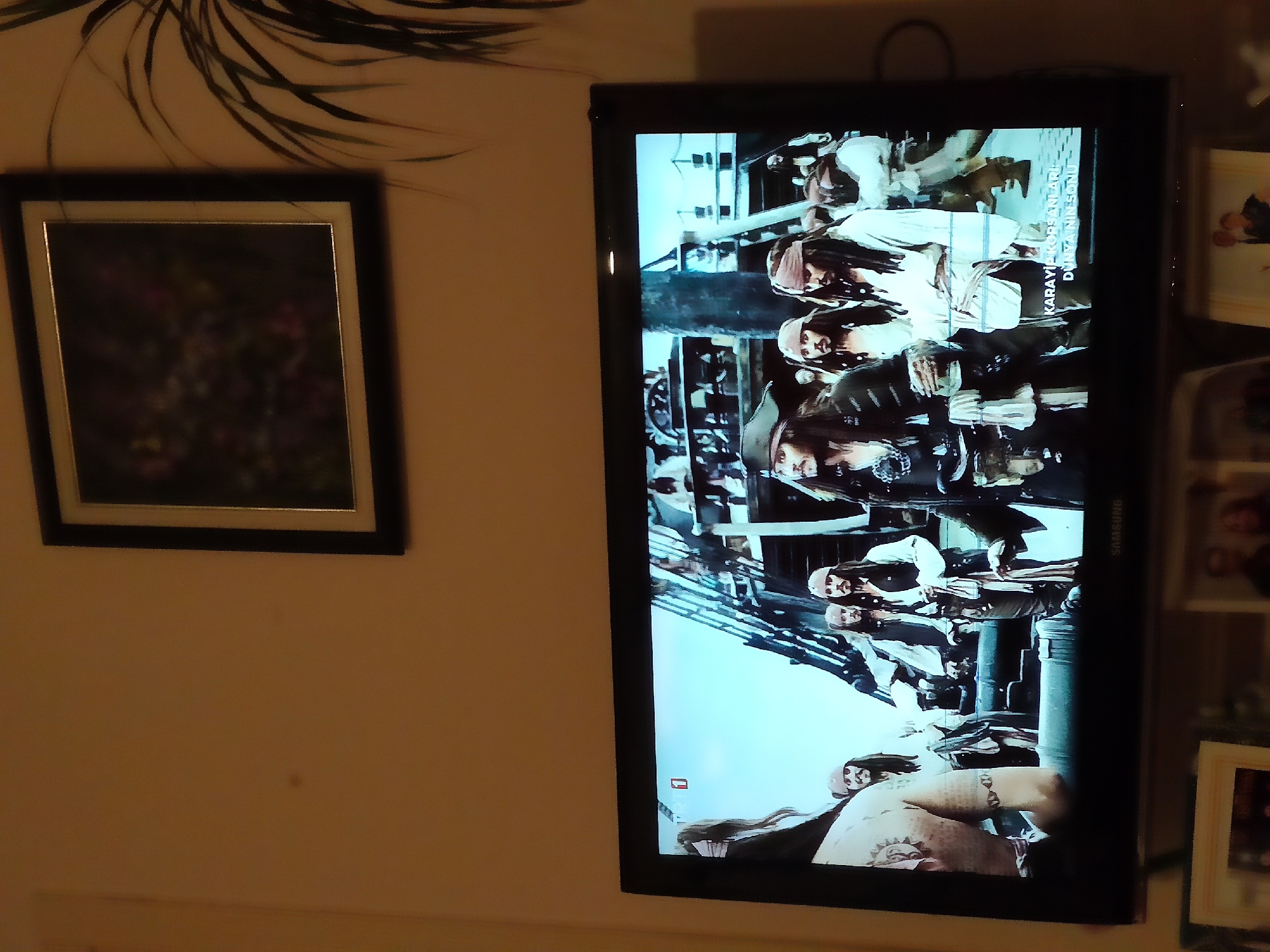 The image shows a television screen displaying a scene from the movie "Pirates of the Caribbean: On Stranger Tides". Several characters, including Captain Jack Sparrow, are visible on a ship's deck. The television is mounted on a wall in what appears to be a living room. In the background, a framed picture hangs on the wall. Below the television, a portion of another framed picture is visible. The scene is dimly lit, suggesting it is nighttime or the room is dark. The television screen shows the movie title in Turkish text: "KARAYİP KORSANLARI DÜNYA NIN SONU", which translates to "Pirates of the Caribbean: End of the World". This scene is captured in a residential setting, likely within the city of Etimesgut, Turkey.