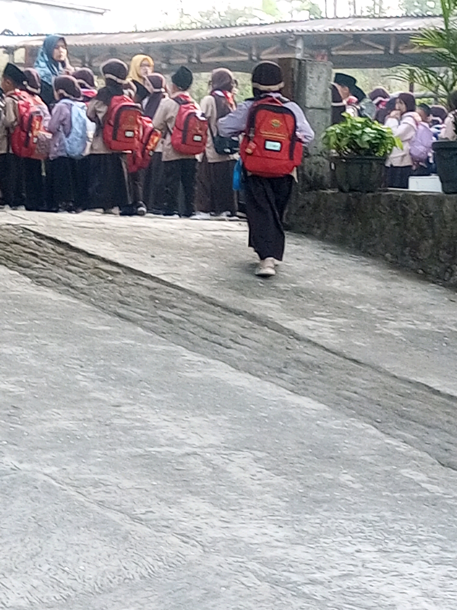 A group of students, dressed in school uniforms and wearing backpacks, are standing in front of a building. One student in the foreground is walking away from the camera, showing the back of their red backpack and dark pants. The students appear to be in an outdoor setting, possibly a schoolyard or a street. The building in the background has a corrugated metal roof and appears to be a simple structure. The ground is paved with concrete. The image quality is low, with a grainy texture and muted colors. The overall scene depicts a casual moment with students gathered outside.