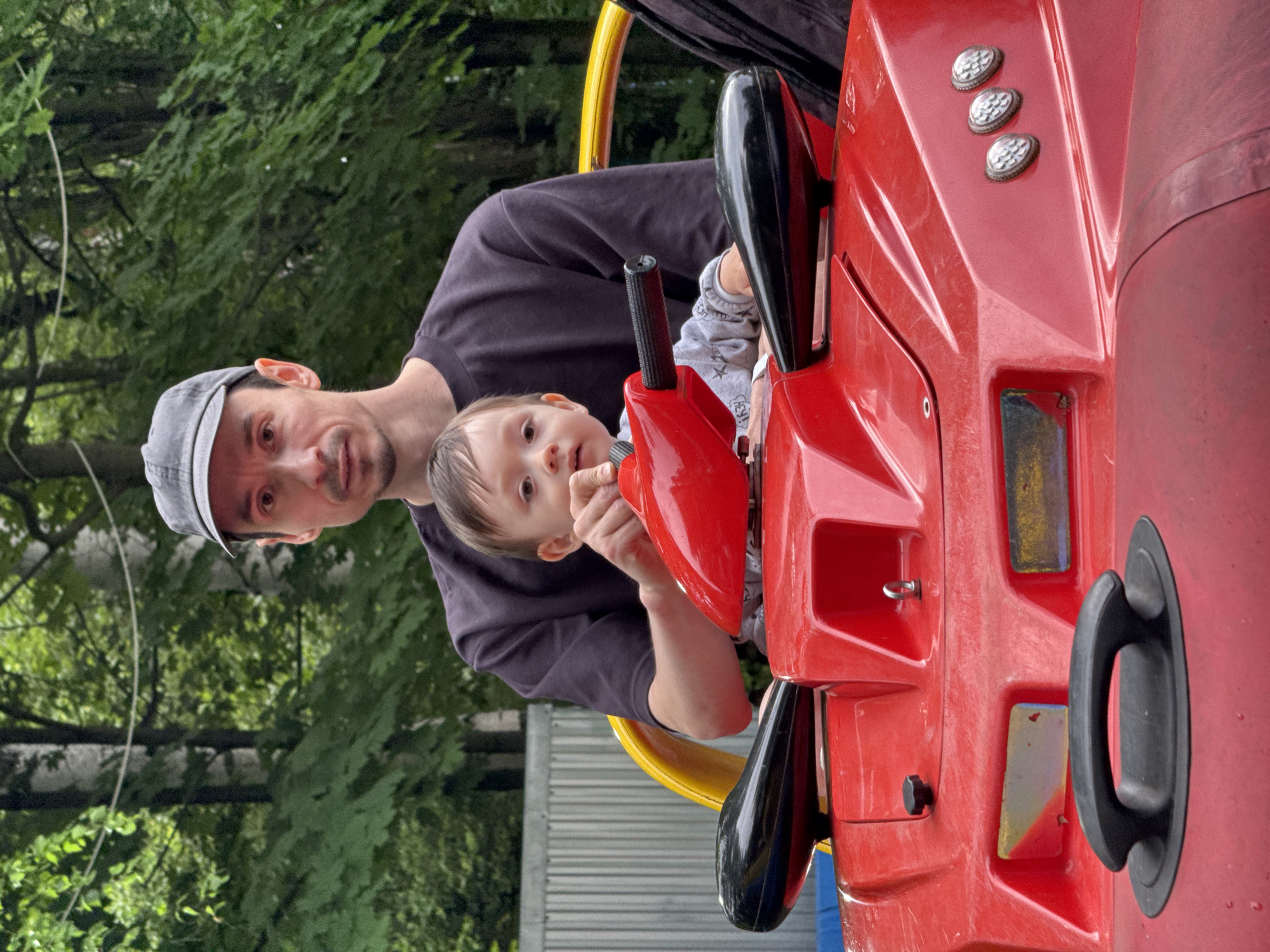 A man in a gray baseball cap and a dark t-shirt is sitting behind a toddler on a red toy ATV. The toddler, wearing a gray long-sleeved shirt, is in the driver's seat and has their hand on the steering wheel. The man's hand is visible, resting on the steering wheel near the toddler's hand. They are on an outdoor playground, with lush green trees and foliage in the background, suggesting a park or a recreational area. A grey metal fence is visible behind the ATV. The scene is captured from a low-angle perspective, focusing on the two individuals and the toy vehicle.