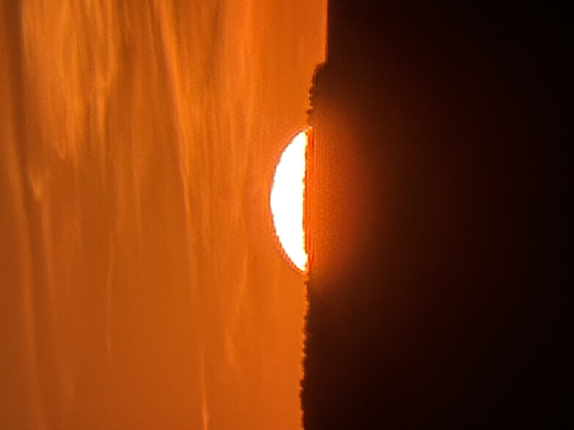 The image depicts a bright, crescent-shaped light source peeking through a vertical gap between two dark orange surfaces. The left surface appears textured, like fabric or drapery, with vertical ripples or folds. The right side is a dark, opaque space. The contrast between the vibrant light and the dark surroundings creates a stark visual effect. There is no indication of a specific location in Chaikovsky, Russia, or any information about people, subjects, objects, actions, or interactions in the image.