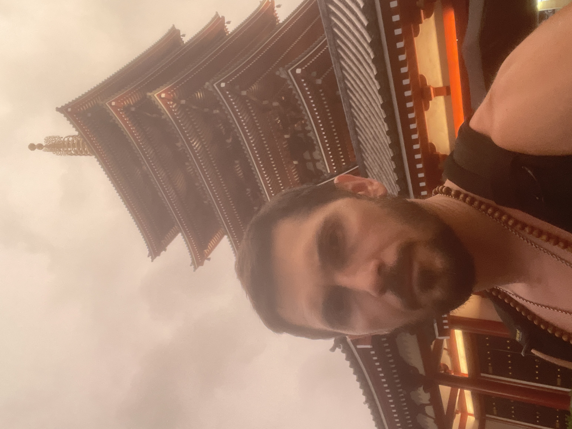 A man is taking a selfie with a pagoda in the background in Taito, Japan. The man is looking directly at the camera with a neutral expression. He has short, dark hair and a beard. He is wearing a black shirt and several necklaces. The pagoda is a tall, five-tiered structure with red and gold accents. The sky is overcast and the overall lighting is dim. The image appears to be taken from a low angle, with the pagoda dominating the background.
