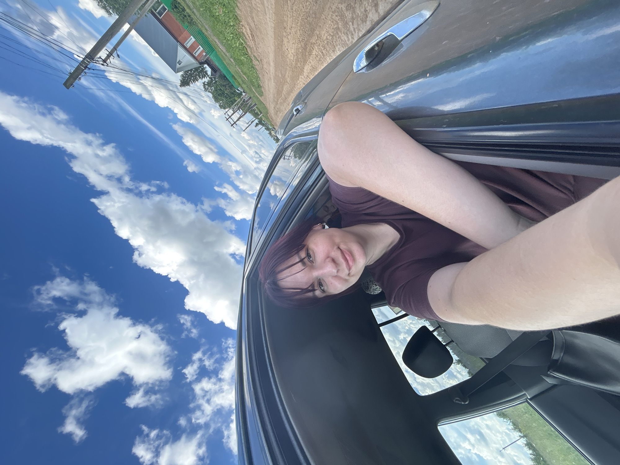 The image shows a young woman with dark hair, wearing a maroon t-shirt, leaning out of a car window. She is holding her arm and hand out of the window, in a selfie pose. The car appears to be on a dirt road, with a rural landscape in the background. There is a house with a red roof and a green fence, utility poles with wires, and trees visible. The sky is blue with white clouds. The image is taken from a slightly low angle, looking up at the woman and the sky. The location is stated to be in the city of Foky, Russia.