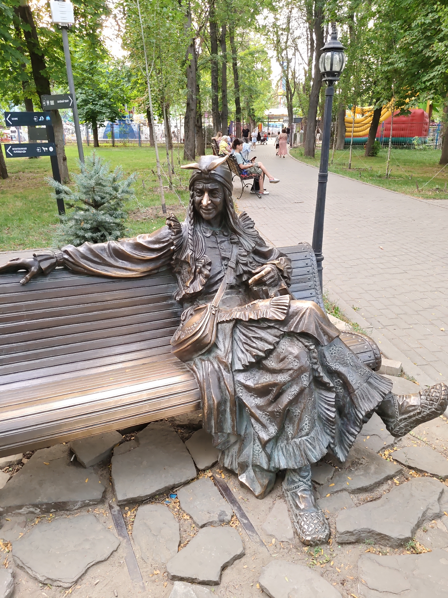 In a park, a bronze sculpture of a person in historical indigenous attire is seated on a slatted park bench. The sculpture is facing slightly to the right and has its left arm resting on the back of the bench, with its right hand extended. The person in the sculpture is wearing a headdress with horns, braided hair, a ruffled collar, and a long, patterned skirt. The sculpture is positioned on a stone-paved area with scattered leaves. Behind the sculpture, a paved walkway winds through a park with numerous trees and some parkgoers walking in the distance. To the left of the sculpture, a directional signpost stands near a small evergreen tree. In the background, a playground with inflatable structures is visible. The overall scene depicts an outdoor recreational area.
