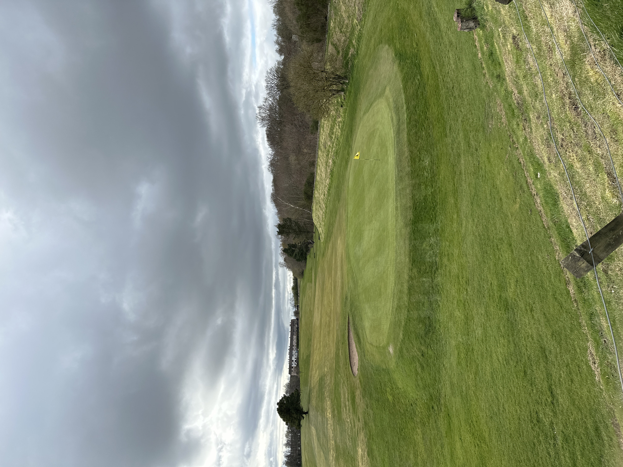The image shows a view of a golf course on a cloudy day. A yellow flag marks the hole on the green. A sand trap is visible on the fairway. A fence lines the right side of the image, with trees marking the boundary beyond the fence. The scene is likely located in Dunfermline, United Kingdom, based on the general location. Beyond the golf course, the sky is overcast with heavy clouds.