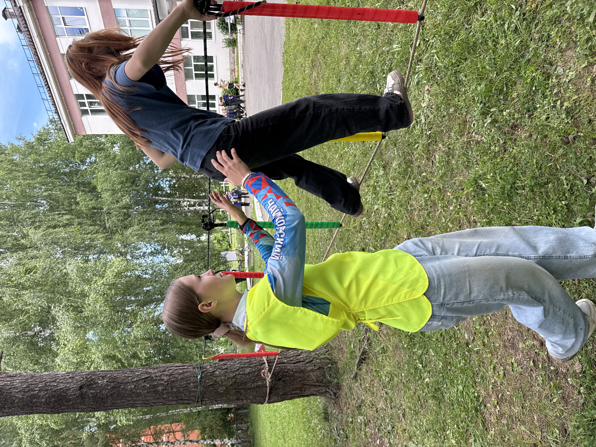 In Chaikovsky, Russia, two teenage girls are participating in an outdoor obstacle course. The girl in the foreground, on the left, wears a bright yellow vest over a blue shirt with colorful graphics and light-wash jeans. She is looking to her right and extending her arms forward, appearing to support or guide the other participant. The second girl, on the right, has long red hair and wears a dark blue t-shirt and dark pants. She is walking on a thick rope strung horizontally between two poles, one red and one yellow. She is leaning forward, with her right arm extended backward for balance and her left hand reaching towards the rope. The environment is grassy with trees and a building in the background, suggesting a park or schoolyard setting.