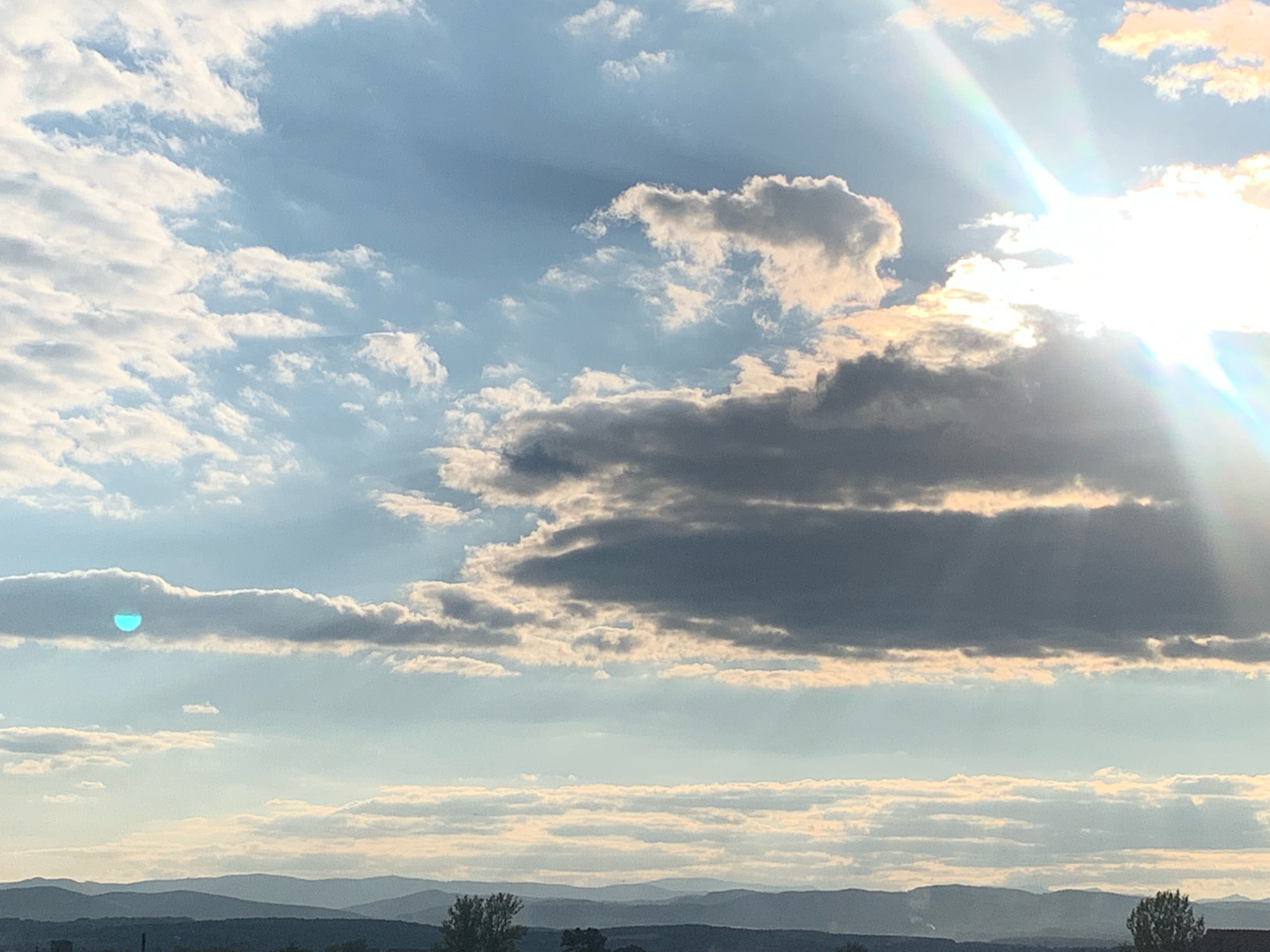 This image depicts a sky filled with a variety of clouds over a distant, hazy landscape of rolling hills. The sun is prominently visible on the right side of the frame, casting bright rays and creating a lens flare effect with a rainbow-like spectrum. The clouds vary in density and color, with some appearing bright white and illuminated by the sun, while others are darker gray, suggesting depth and possibly an approaching weather system.
A distinct, bright turquoise circular or semi-circular shape is visible within a band of clouds on the left side of the image. The lower portion of the image shows a silhouetted horizon line with layered hills and a dark foreground that appears to be a city or town with some vegetation. The sky occupies the majority of the frame. There are no discernible people, actions, or interactions in this image. The location within Kolomyia, Ukraine, is not visually identifiable from the provided image alone.