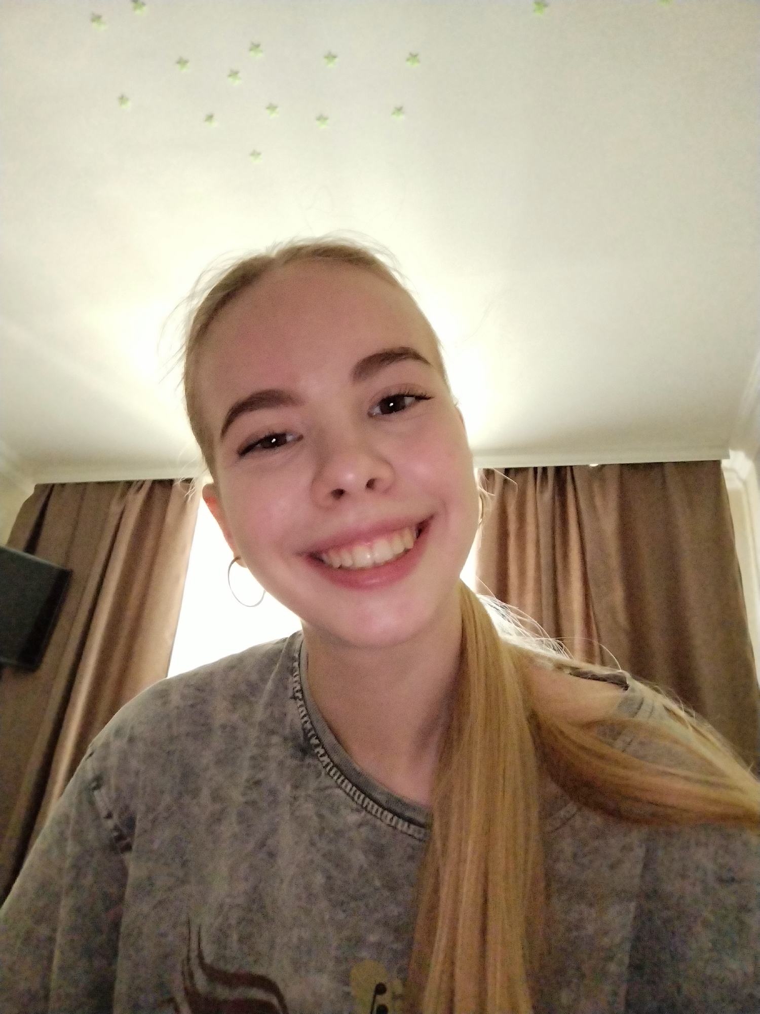 A young woman with blonde hair pulled to the side is taking a selfie indoors. She is smiling widely and her head is tilted. She is wearing a grey, textured t-shirt. The background consists of a white ceiling with small, green star-shaped glow-in-the-dark stickers, and brown curtains. A dark object, possibly a screen or monitor, is partially visible to the left of the woman. The lighting suggests an indoor setting, possibly a bedroom.
