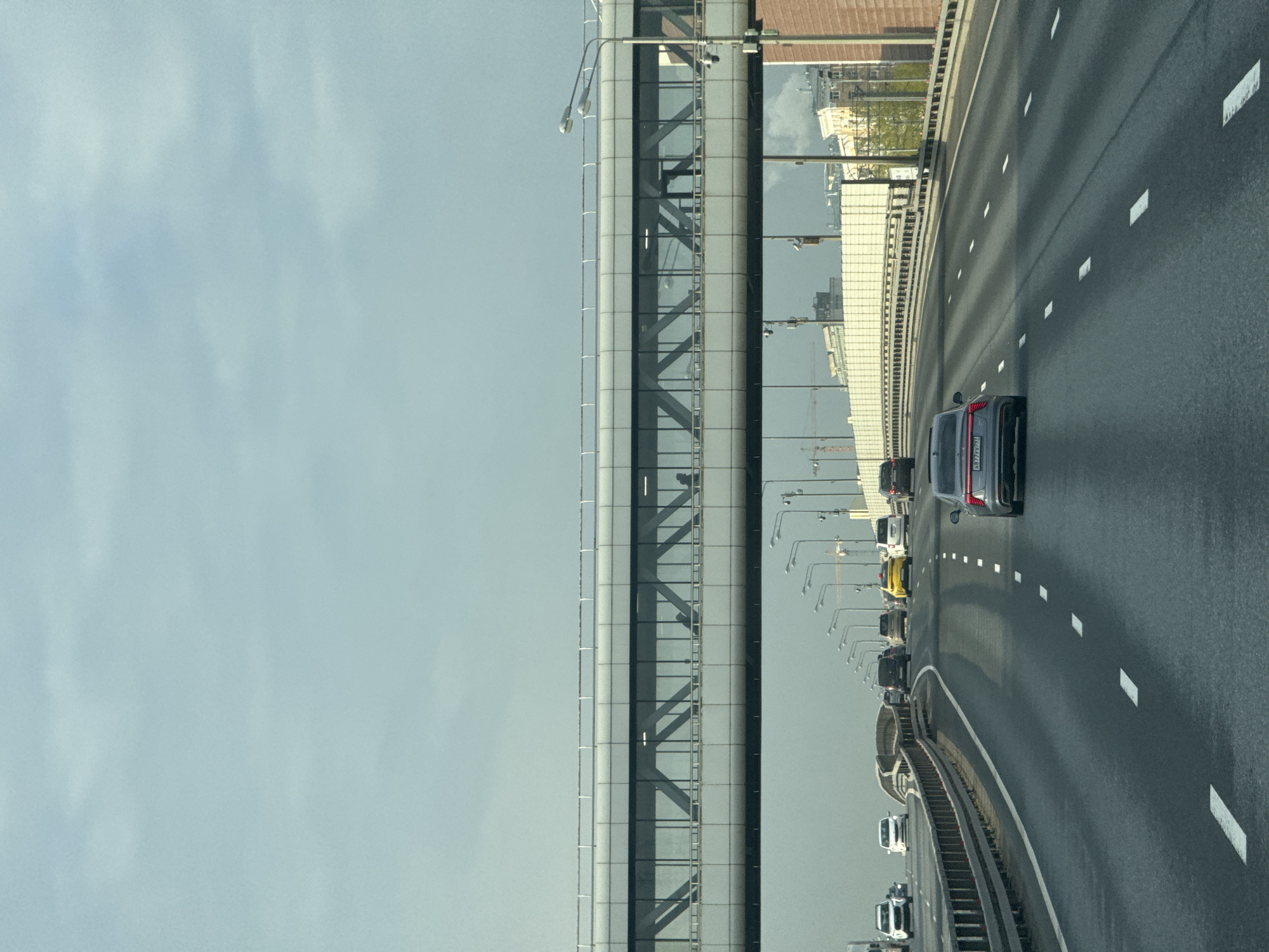 The image depicts a multi-lane highway in Moscow, Russia, oriented vertically. The highway is densely packed with cars traveling in the same direction, with a grey Audi SUV being the most prominent vehicle visible in the right lane. The road surface is wet, reflecting light. White lane markers are visible on the road. A modern overpass or bridge structure runs parallel to the highway on the left side of the image. Lampposts line the side of the highway. The sky is overcast. Buildings are reflected in the glass portions of the overpass. A construction crane is visible in the distance. The license plate of the SUV reads "A917YX797".