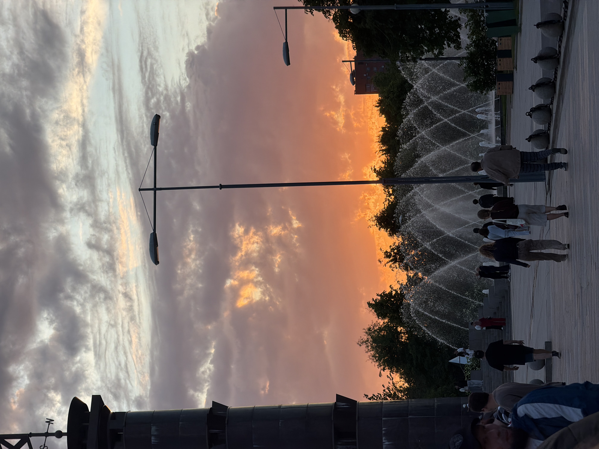 This image captures a vibrant sunset scene in a public park or promenade in Saint Petersburg, Russia. The sky dominates the upper portion, displaying a dramatic gradient from intense orange, pink, and yellow hues on the right, where the sun is setting, to softer gray and white clouds on the left.
In the mid-ground, a series of decorative fountains create an intricate pattern of crisscrossing water jets, illuminated by the fading light. A wide, paved pathway runs alongside these fountains, where numerous people are seen casually strolling, standing, or observing the beautiful surroundings. Many individuals are partially silhouetted against the bright sky. Dark, lush trees line the perimeter behind the fountains, with a multi-story building visible through their branches in the distance.
Several tall, modern streetlights with oval lamps are positioned throughout the area. A distinctive row of large, round stone bollards, connected by heavy chains, separates the pathway from the fountain area. In the foreground, at the bottom left, a section of a large, dark, architectural or sculptural structure is visible. This structure includes a weather vane with the cardinal directions 'N' and 'W' clearly marked.
The scene conveys a peaceful and leisurely atmosphere, characteristic of an evening gathering in a public recreational space, greatly enhanced by the spectacular sunset.