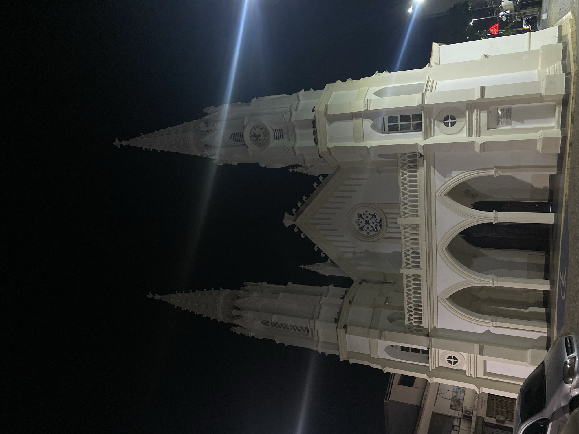 The image depicts the Iglesia San Juan Evangelista (Church of Saint John the Evangelist), located in Juan Griego, Venezuela, at night. The white facade of the church is prominently displayed, featuring two towers with spires, a clock on one of the towers, arched doorways, and decorative windows. The sky is dark. A streetlight is visible above the church, casting a bright glow. A vehicle is partially visible on the right. The overall scene suggests a quiet night in the city.