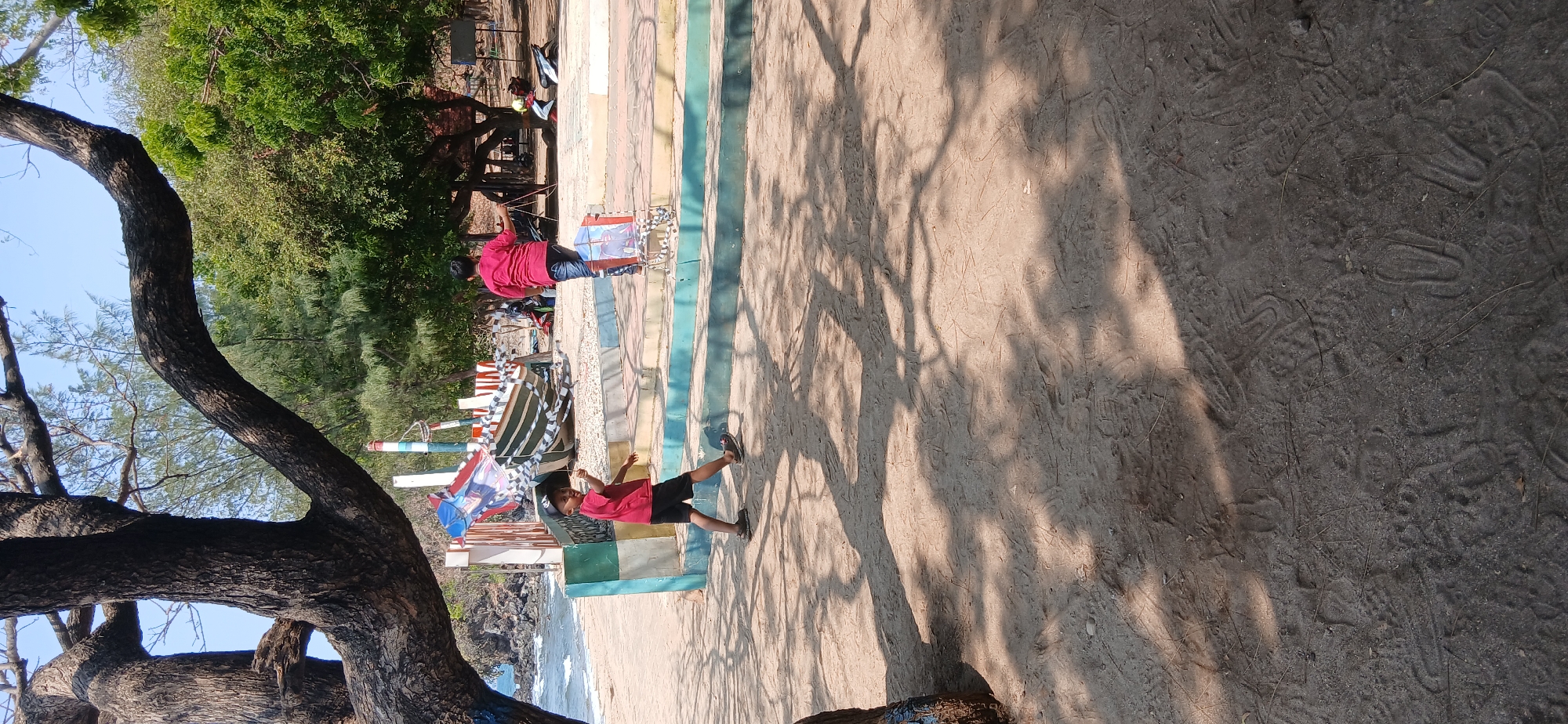 In Banyuglugur, Indonesia, two boys are playing on a sandy beach near a large tree. One boy, wearing a red shirt and black shorts, is holding a kite and appears to be running. The other boy, in a red shirt and dark pants, is walking away from the camera. In the background, a partially constructed boat is visible, along with trees and the ocean. The ground is covered in sand with visible footprints and tree shadows.