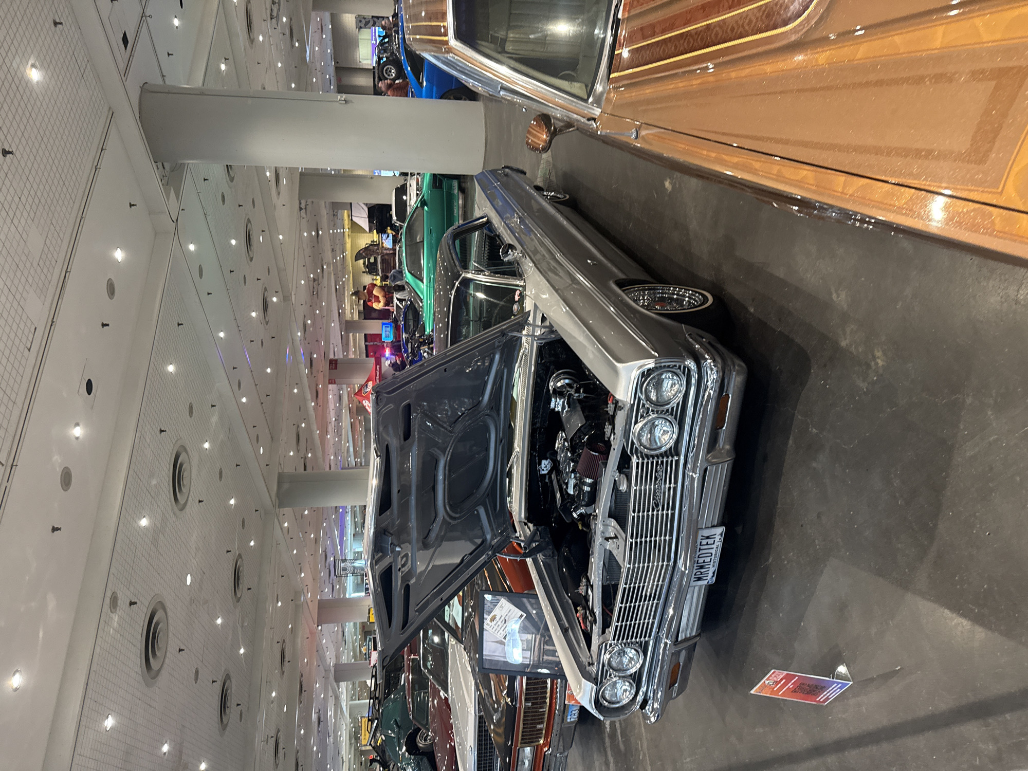 The image shows an indoor car exhibition, likely a custom car show, in a city and specific location marked as Unknown, Unknown. The primary subject is a silver classic car with its hood open, revealing the engine. A decorative license plate is visible on the front of the car. Beside the silver car is a bronze-colored car. Other cars, including a green car, are partially visible further in the background. The exhibition space is large with a high ceiling that includes rows of lights and ventilation ducts. There are pillars providing structural support within the space. No people are clearly visible in the foreground. A small promotional sign is on the floor near the silver car.