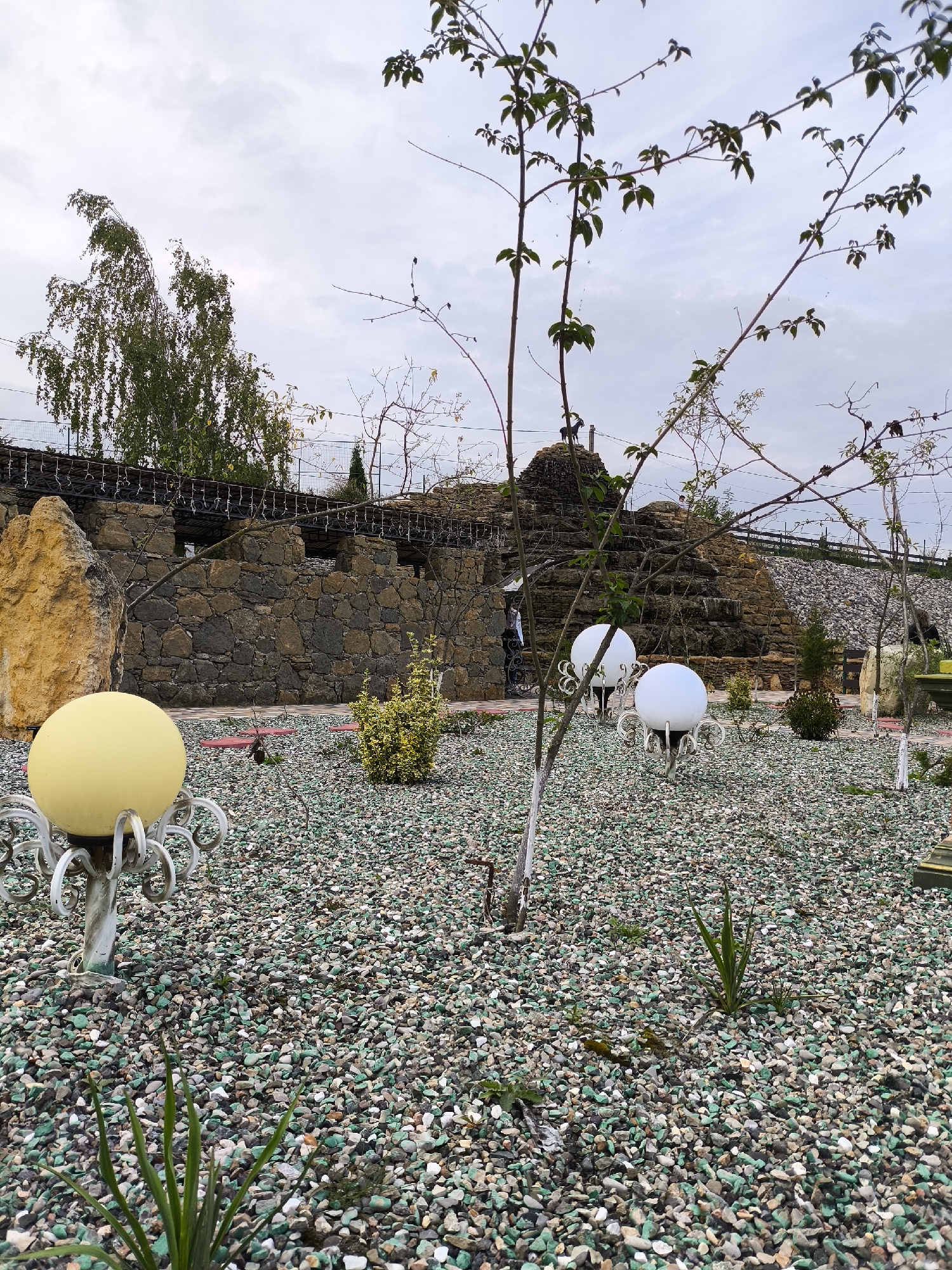The image shows a park area with a rocky landscape and greenery. In the foreground, a pale yellow spherical lamp is visible, mounted on a decorative metal stand. The ground is covered with a mix of small, multi-colored pebbles, predominantly green, white, and brown. Several young trees with bare branches and a few leaves are scattered throughout the scene.
In the middle ground, two white spherical lamps with similar decorative stands are positioned. Behind the lamps, a tiered rock structure resembling a waterfall or a stepped hill is visible, constructed from rough-hewn stones. A stone wall with arched openings forms a backdrop to this structure. A person in dark clothing is partially visible near the rock structure.
The background features more trees, some with sparse foliage, and a cloudy sky. A fence with a wire mesh is present along the top of the stone wall and extends into the background. The overall impression is that of an outdoor recreational space with an emphasis on natural and decorative elements. The specific location in the city of Borgustanskaya, Russia, is not visually indicated in the image.
