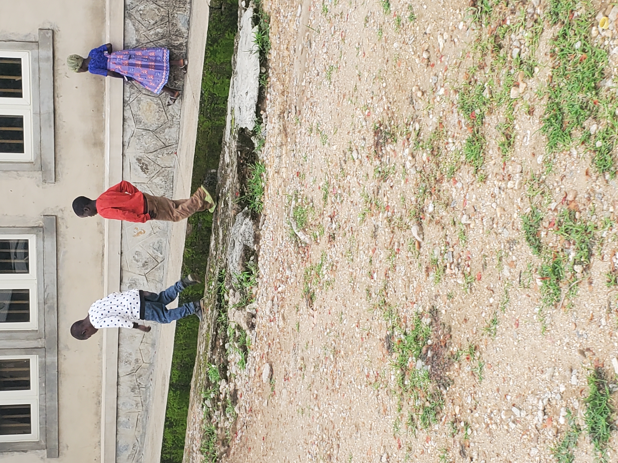The image depicts an outdoor scene with three children walking along a pathway next to a building. In the foreground, the ground is covered with gravel and sparse green vegetation. The middle ground features a raised concrete pathway with mossy edges, separating the viewer from the building. The building in the background has a beige facade with large, horizontally arranged windows.
The child furthest to the left is wearing a white long-sleeved shirt with black polka dots and blue jeans. They are walking away from the viewer, looking down. The middle child is wearing a red long-sleeved shirt and brown pants. They are also walking away from the viewer, with their hands in their pockets. The child furthest to the right is a girl wearing a blue and purple patterned dress and a headscarf. She is walking away from the viewer and looking back over her shoulder. The children appear to be walking in the same direction along the pathway. The overall environment suggests an urban or semi-urban setting with some overgrowth.