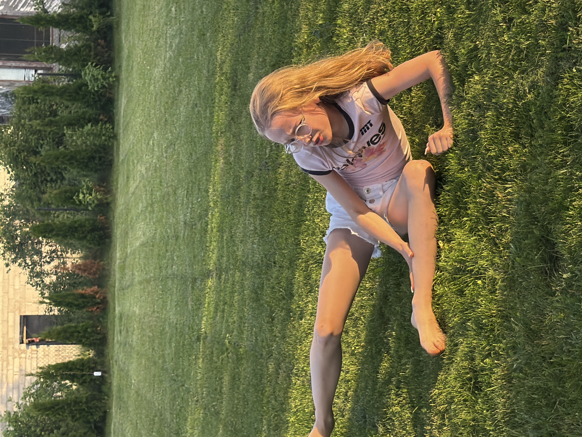 A young blonde girl with long hair and glasses sits on a vibrant green lawn. She is wearing a light pink t-shirt with black trim and white shorts. The t-shirt has a graphic print with visible text. She is leaning on her right arm, with her left leg bent and her right leg extended. Her left hand is touching her right shin. The grass is well-maintained and appears to be illuminated by the warm glow of what seems to be late afternoon or early evening sunlight. In the background, there are several evergreen trees and a building with a pale, brick-like facade and a dark window. The overall scene suggests a relaxed outdoor setting, possibly a backyard or park, in Dreiliņi, Latvia. The lighting and the girl's posture convey a sense of ease and comfort in the outdoor environment. The image is oriented vertically, with the girl and the grass filling most of the frame.