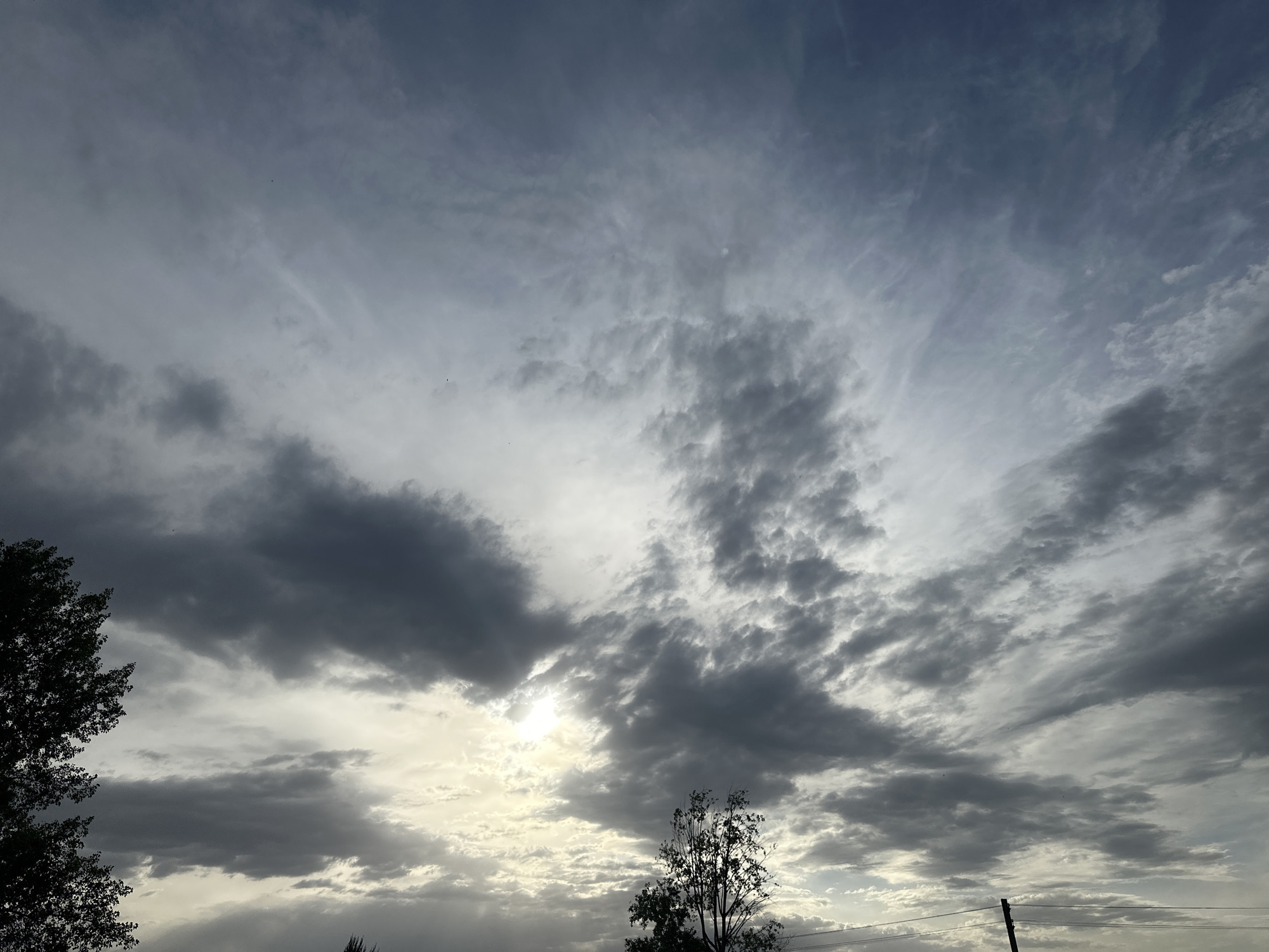 The image captures a sky dominated by varying cloud formations. The clouds range from dense, dark gray masses to thinner, wispy, white streaking across the upper portion of the frame. Sunlight filters through the clouds, creating a diffused, bright area in the lower-center.
Silhouetted trees are visible on the left and in the lower-center. A power pole is visible at the bottom right of the image. The image provides no evidence of people or actions. The location in the city of Chaikovsky, Russia, cannot be confirmed from the information provided.