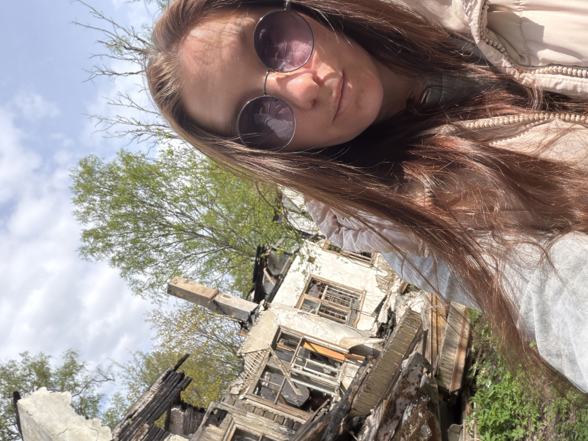 A selfie taken outdoors, likely in the city of Tokkari, Russia, given the query context. The subject is a woman with long, brown hair and wearing round, tinted sunglasses. She is also wearing a light-colored jacket or coat. Behind her is the ruined remains of a building, appearing to be heavily damaged, possibly by fire. Some charred wooden beams and debris are visible, along with parts of the white walls. Green trees and a cloudy sky are visible in the background above the destroyed building.