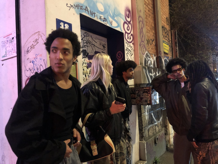 The image depicts five individuals gathered at night on an urban street in Bagnolet, France. The scene is illuminated by artificial light sources, casting pinkish and yellowish hues. A building wall, heavily covered in graffiti, dominates the background.
From left to right:
1. A male with dark, curly hair and light brown skin is positioned in the foreground, wearing a black long-sleeved shirt, an open black jacket, and light-colored pants. He is looking intently towards the left of the frame, with his right hand near his waist.
2. A female with blonde, shoulder-length hair and light skin is positioned slightly behind the first male, wearing a black puffer jacket and carrying a light-colored shoulder bag with a prominent gold-colored buckle. She holds a dark smartphone in her right hand, looking downwards.
3. Partially obscured behind the female is a male with dark, afro-style hair and dark skin, wearing a dark jacket and camouflage-patterned pants. He is looking towards the right side of the frame.
4. To the right, another male with dark, afro-style hair and dark skin is visible, wearing orange-tinted sunglasses, an olive-green jacket, and dark pants. He is leaning against the graffiti-covered wall, resting his right arm/hand near his head, and looking towards the right.
5. On the far right, a fifth male with dark dreadlocks and dark skin is partially visible, wearing a black leather-like jacket and camouflage-patterned pants. He is facing the fourth male, appearing to be engaged in conversation.
The building wall behind them is composed of brick on the right, transitioning to a smoother, graffiti-covered surface on the left. Above a dark doorway, a blue sign with the number "12" is visible. The wall also features visible graffiti including "SAMPLE-ANTIFA" above the doorway. A white poster with diagrams is affixed to a wall section on the far left, bathed in pink light. Bare tree branches are visible in the upper right corner against the night sky.