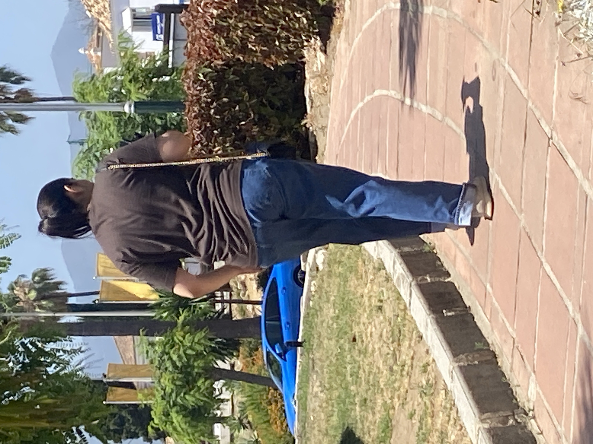 A person is walking away from the camera on a red tiled pathway. The person is wearing a dark brown t-shirt, blue jeans, and light-colored shoes. They have dark hair pulled back in a ponytail and are carrying a purse with a gold chain strap over their right shoulder. The pathway curves to the right and is bordered by a low stone wall and green grass. In the background, a bright blue car is parked on a patch of grass, partially obscured by the wall and grass. Behind the car are trees and vegetation. Further in the background, there are buildings with white walls and tiled roofs, and palm trees. In the far distance, hills are visible against a blue sky. The scene appears to be outdoors in a sunny, possibly Mediterranean, environment. The location is identified as Resinera-voladilla, Spain.