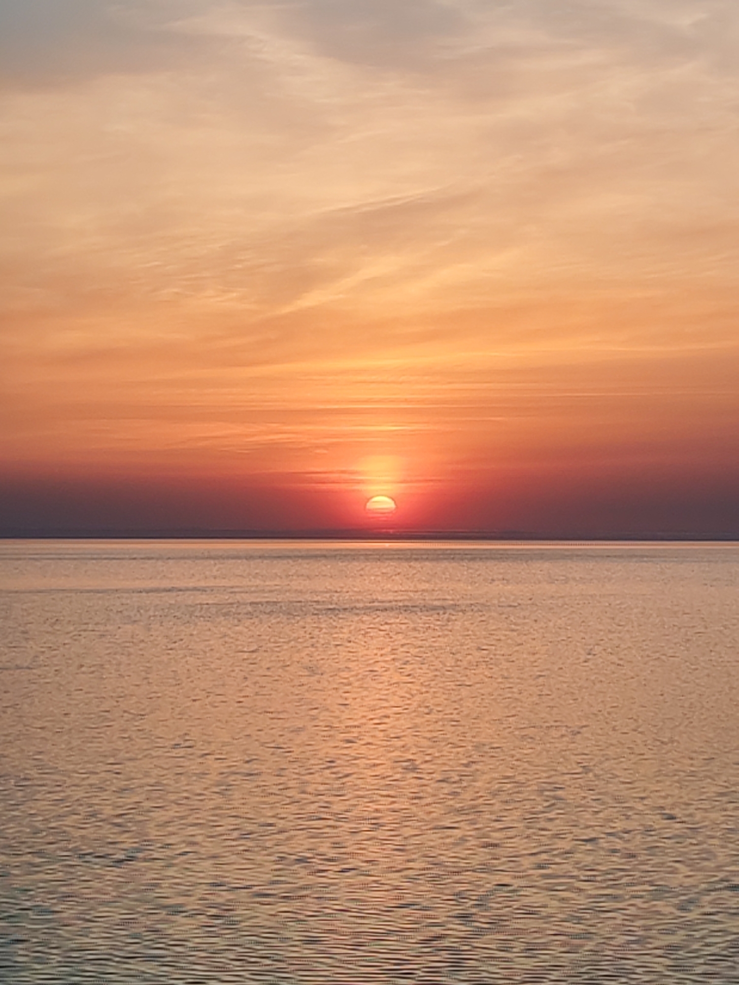 The image shows a sunset over water. The sky is filled with orange and red hues, with the sun partially visible just above the horizon. The water reflects the colors of the sky.