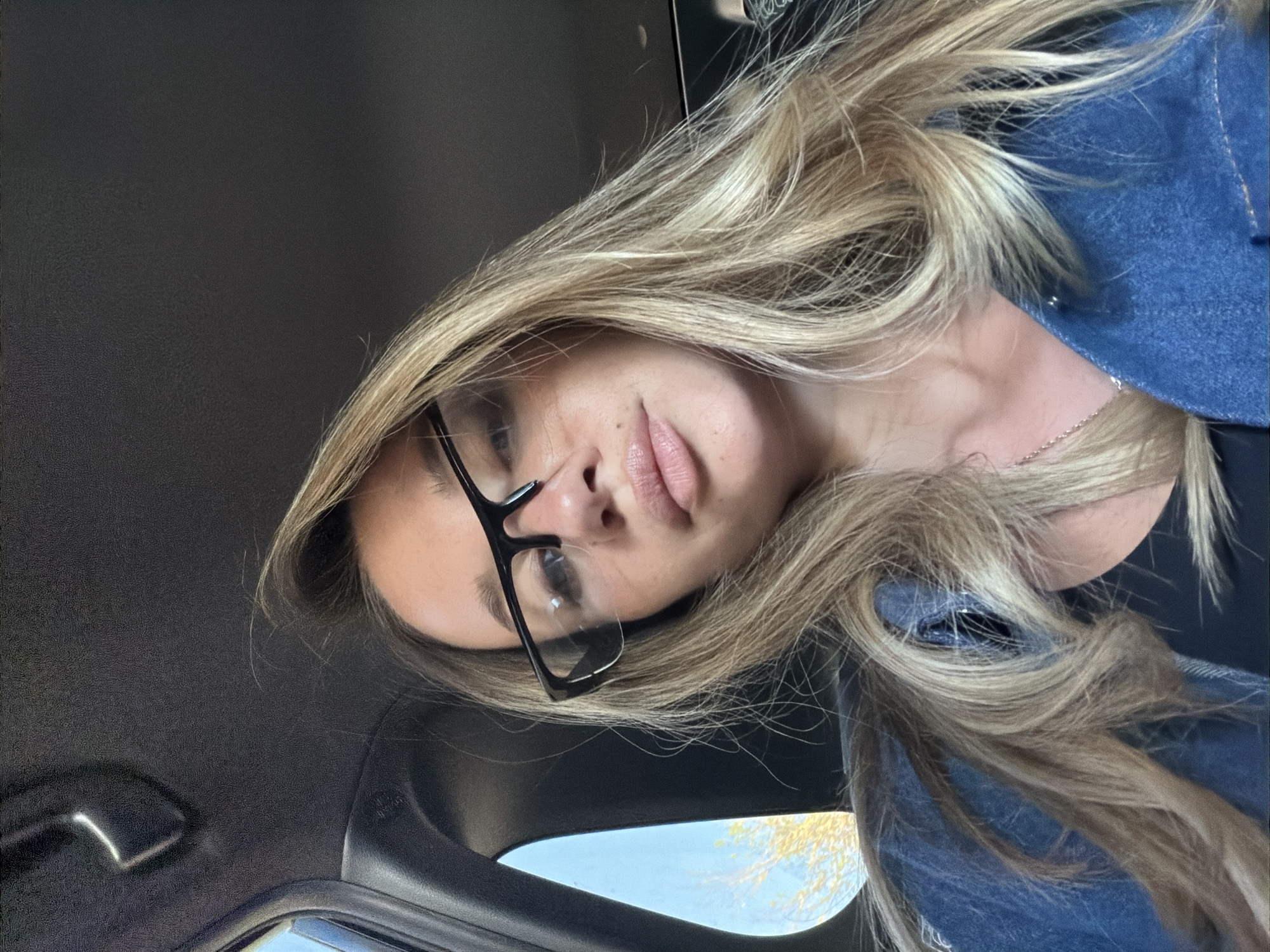 The image shows a woman with blonde hair wearing black glasses and a blue denim jacket. She is positioned in the interior of a car, with the black interior roof visible above and a glimpse of a blue sky and tree branches seen through the car's window. Her face is the primary focus, and she appears to be looking at the camera. The location is in the city of Unknown, Unknown.