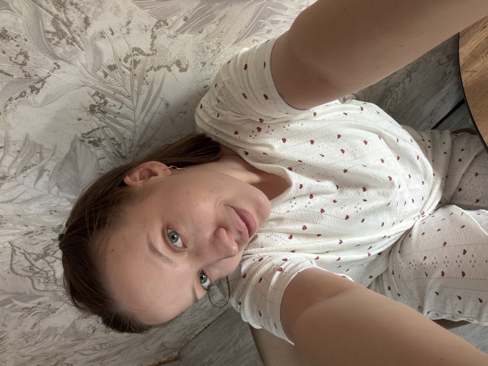 A woman with light skin, brown hair, and blue eyes is taking a selfie indoors. She is wearing a white garment with red heart patterns. She is positioned against a patterned wall. Based on the metadata, this photograph was taken in Reshetnikovo, Russia.