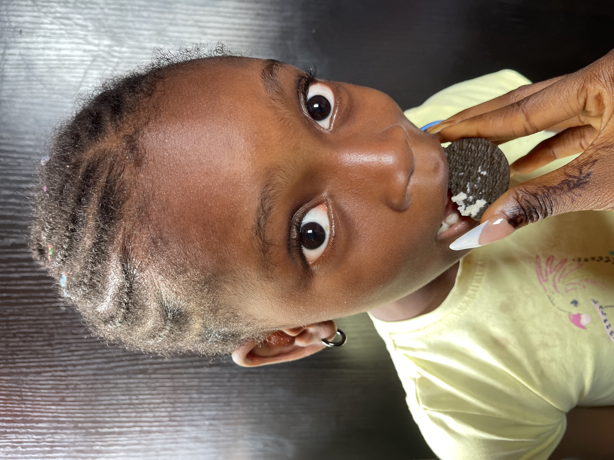 A close-up portrait depicts a young Black female child, estimated to be between 5-8 years old, looking directly at the viewer with wide eyes. Her dark hair is styled in cornrows, some adorned with small colorful beads. She wears a light yellow t-shirt and a silver-colored hoop earring in her left ear. Her mouth is open, with teeth visible, as she consumes a dark-colored sandwich cookie with white cream filling. The cookie is presented to her by a dark-skinned hand, presumably an adult's, characterized by long, pointed acrylic nails with white French tips. The background is a dark, vertically textured surface. The scene is situated in the city of Ado, Nigeria.